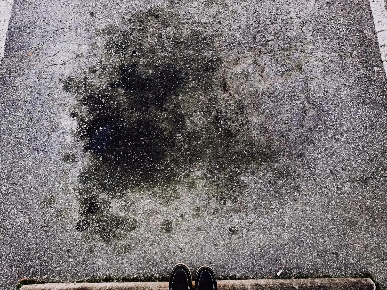 Overhead view of a large dark oil stain soaked into a cracked asphalt parking lot surface, with a person's feet visible near the curb for scale.
