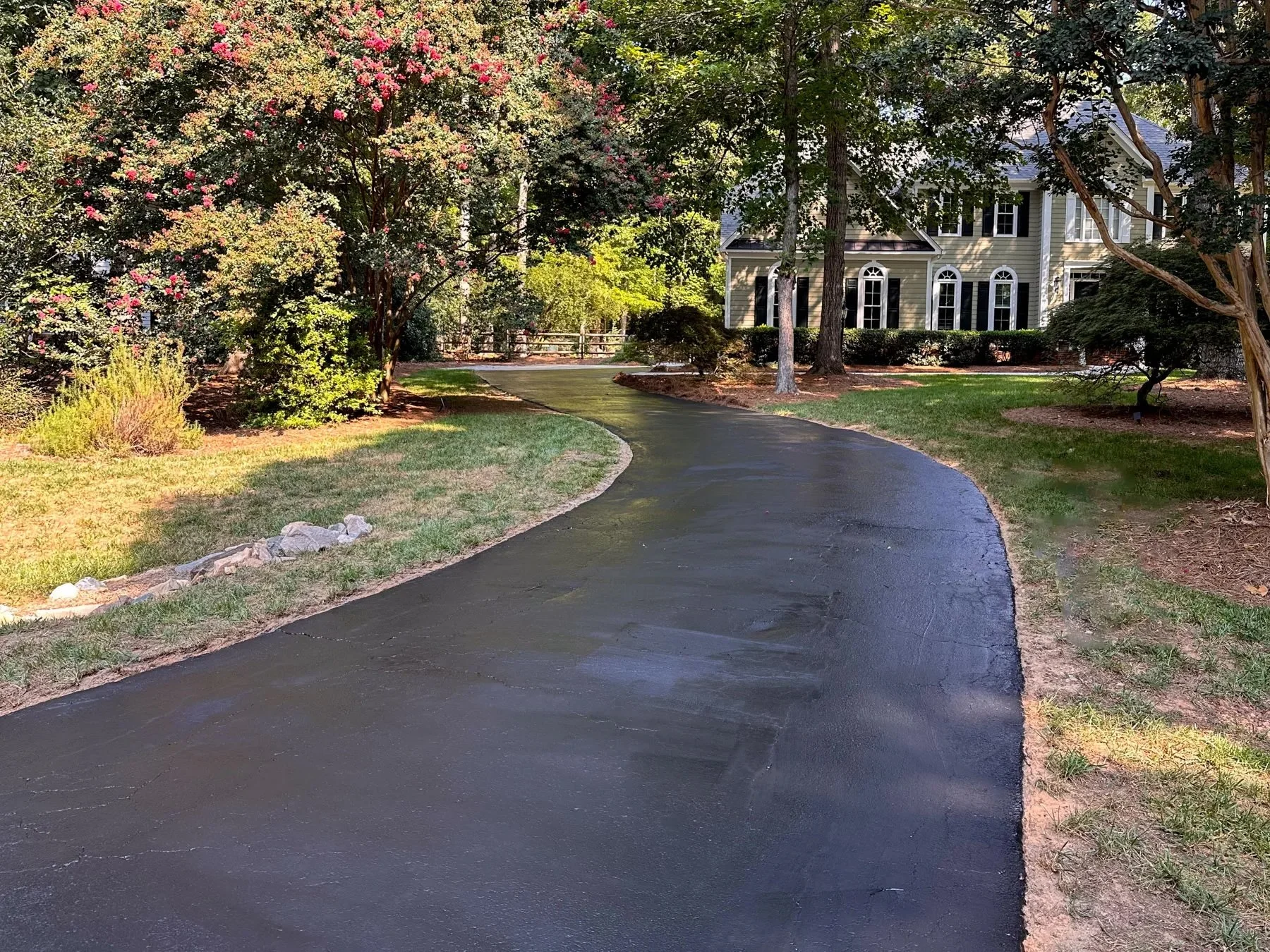 A freshly sealcoated driveway with a smooth, dark black finish winding through wooded, crepe-myrtle-lined landscaping to a cream two-story home in Raleigh, NC.