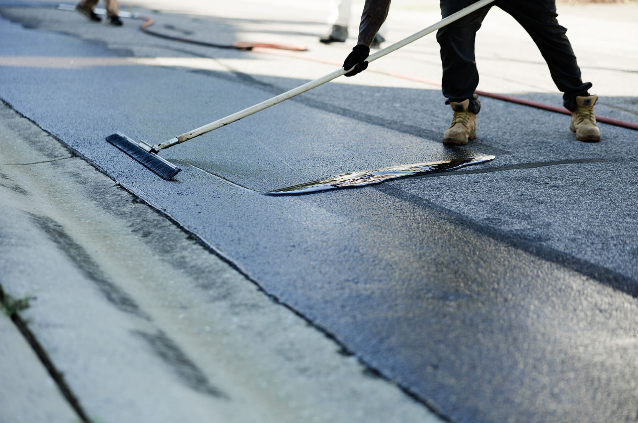 Green Street Blacktop worker applying fresh asphalt sealer with a long-handled squeegee, while another person is visible in the background.