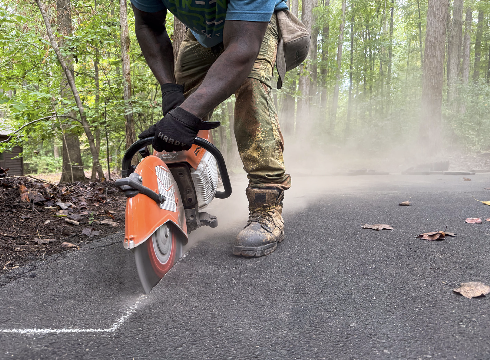 A Green Street Blacktop worker in long pants, boots, gloves, and a shirt is using a handheld power saw to cut through asphalt on a paved road in Durham, NC.