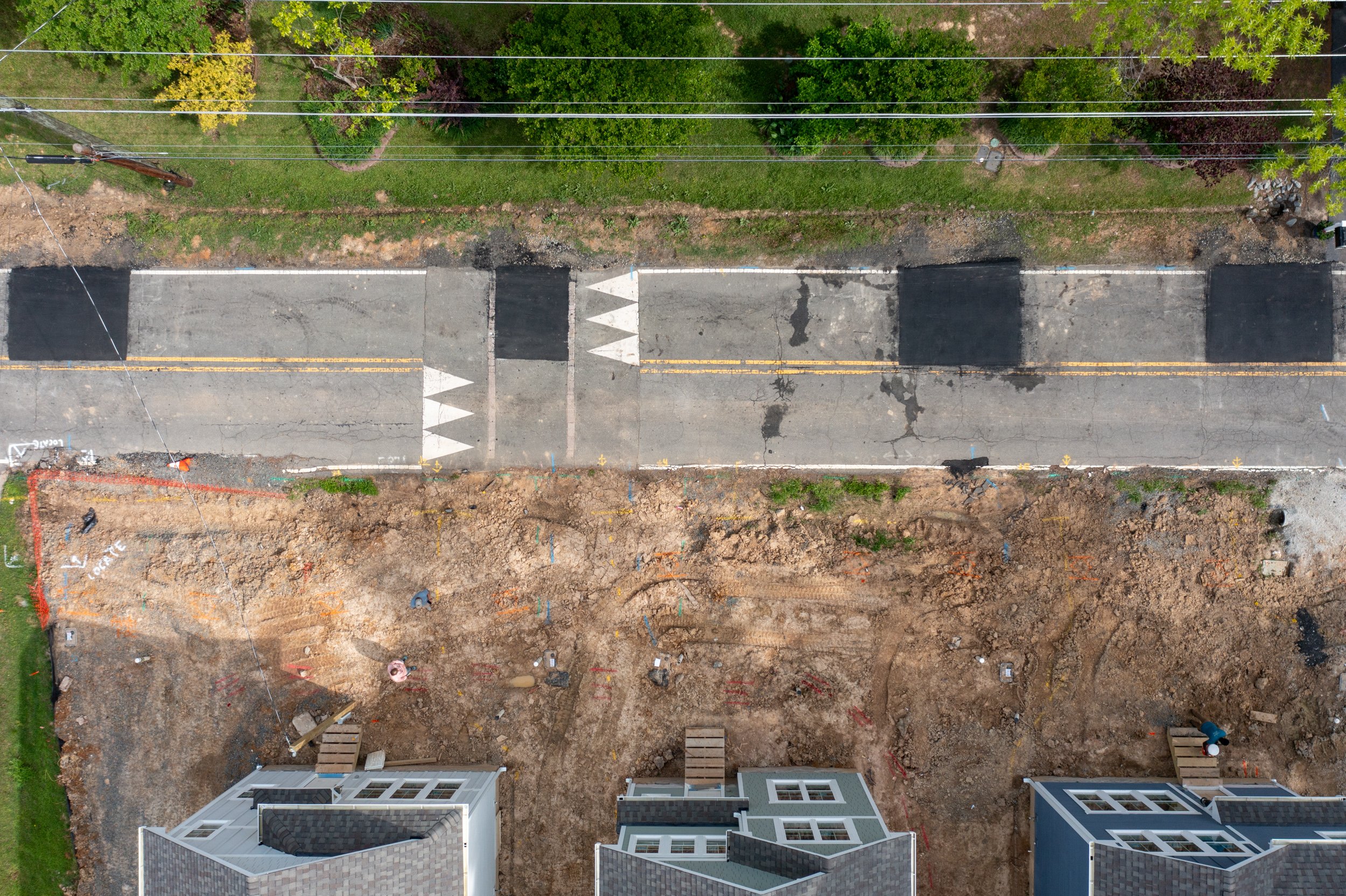 An aerial view of a street under construction with partially paved road and construction site, residential buildings at the bottom, and trees along the top.