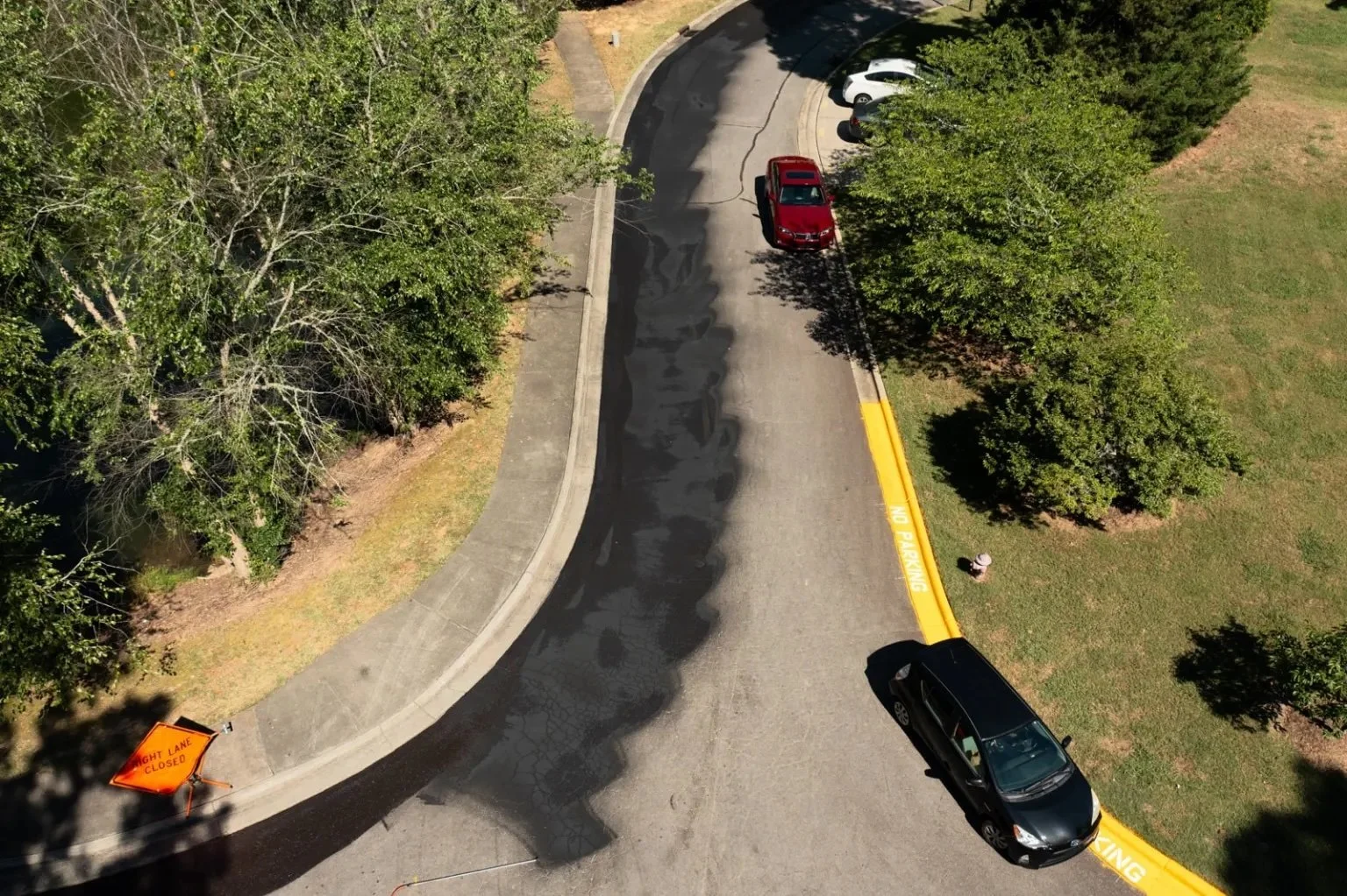 Aerial view of a curving neighborhood road where dark, freshly applied asphalt emulsion covers part of the lane, contrasting with the lighter gray pavement beside it.