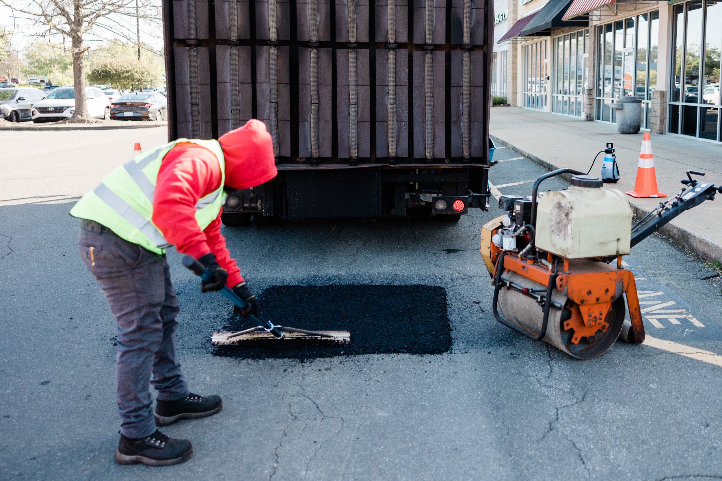 Green Street Blacktop worker in red hoodie and yellow safety vest operating a machine to repair asphalt in a parking lot, with a infrared truck and cars in the background.