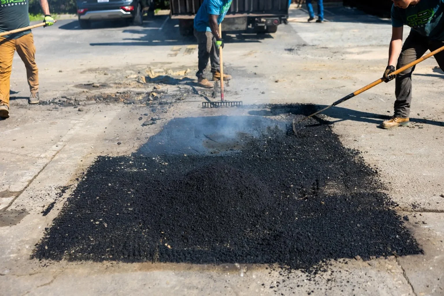 Green Street Blacktop crew members raking fresh hot mix asphalt into a repaired pothole, steam rising from the material, Durham NC parking lot