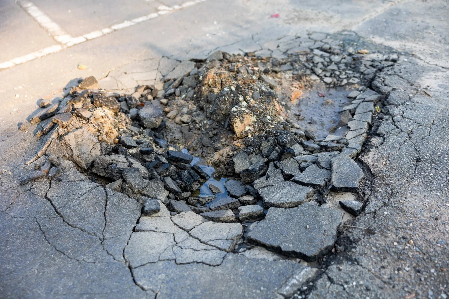 A deteriorated stretch of asphalt pavement with crumbled surface material, exposed cracks, and a pothole holding pooled water.