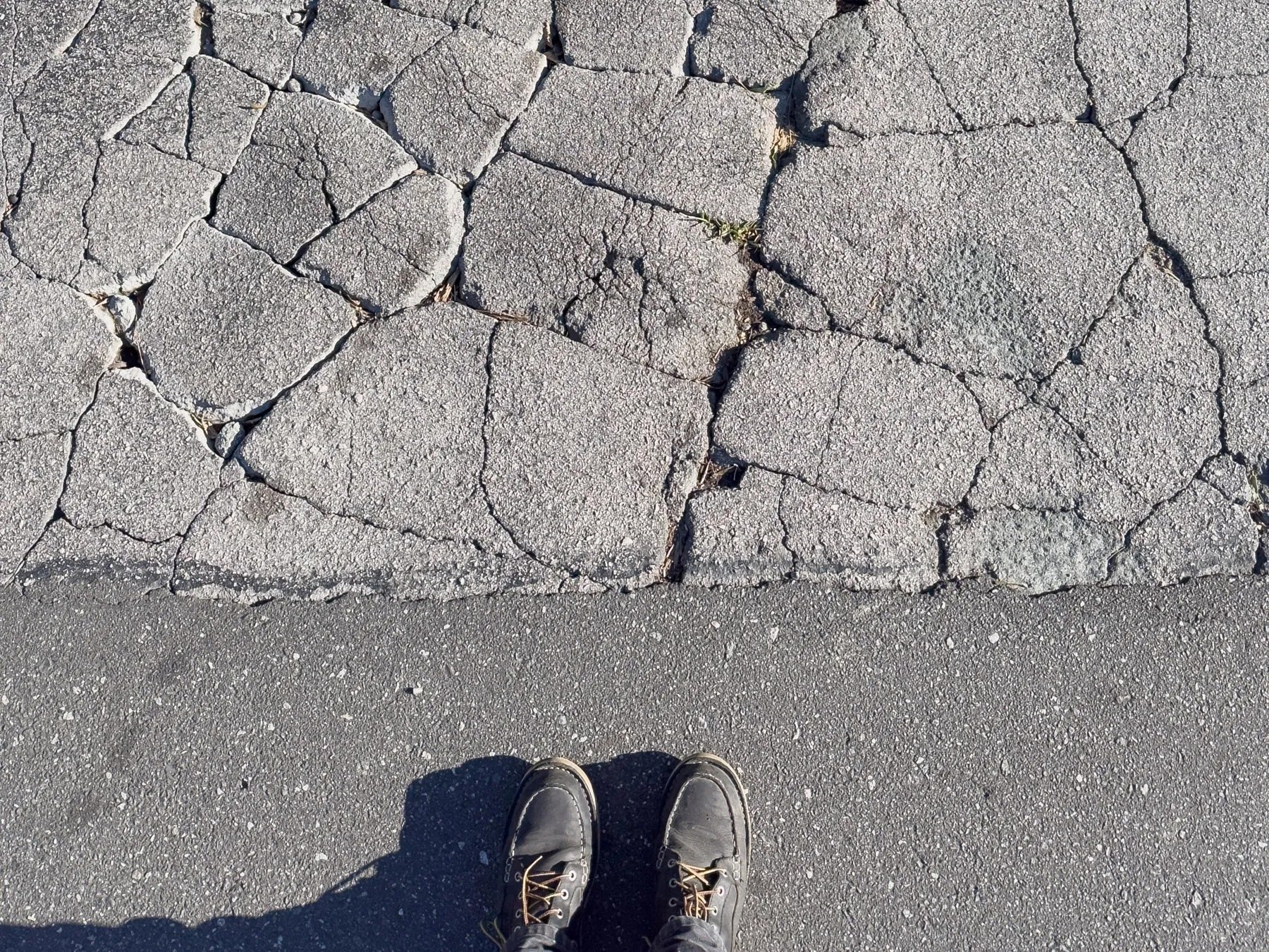 Aerial view of severely cracked and alligatored asphalt pavement, with a person's shoes visible at the edge of the damaged area.