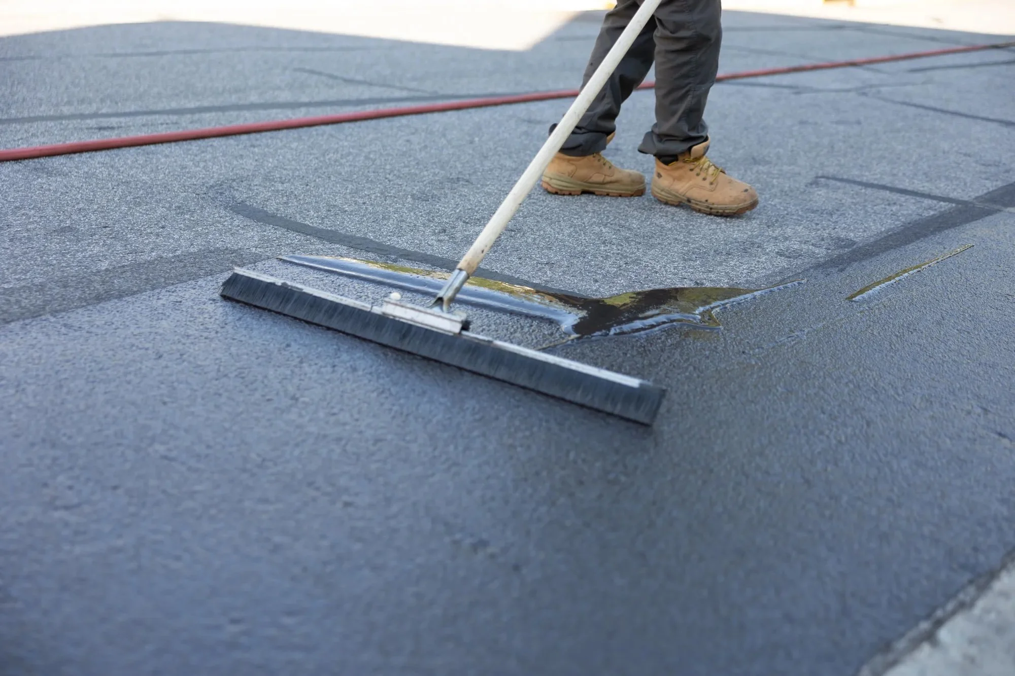 A worker uses a long-handled squeegee brush to spread fresh black sealcoat across a paved surface, with a hose visible in the background