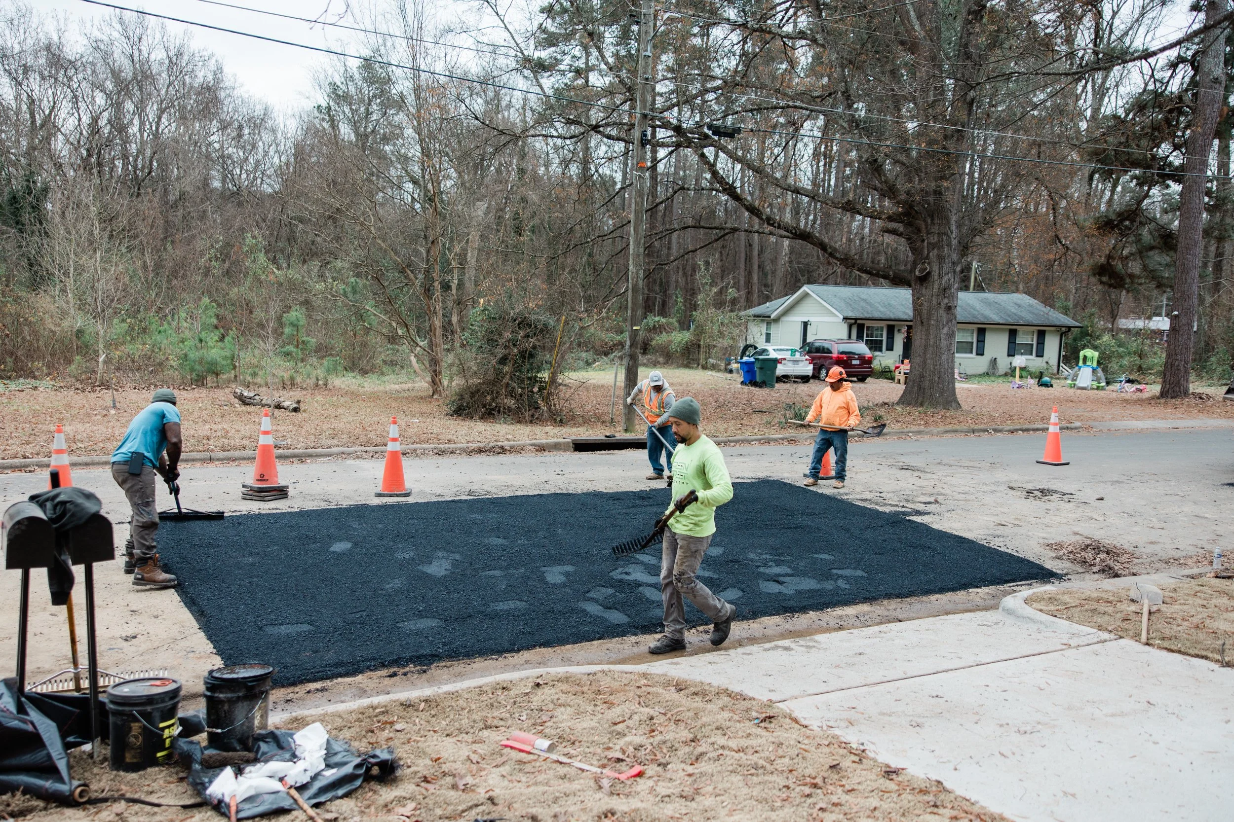 Workers repairing a utility cut with fresh asphalt on a residential street in Durham, NC, wearing safety gear, with houses, trees, and parked cars visible in the background.