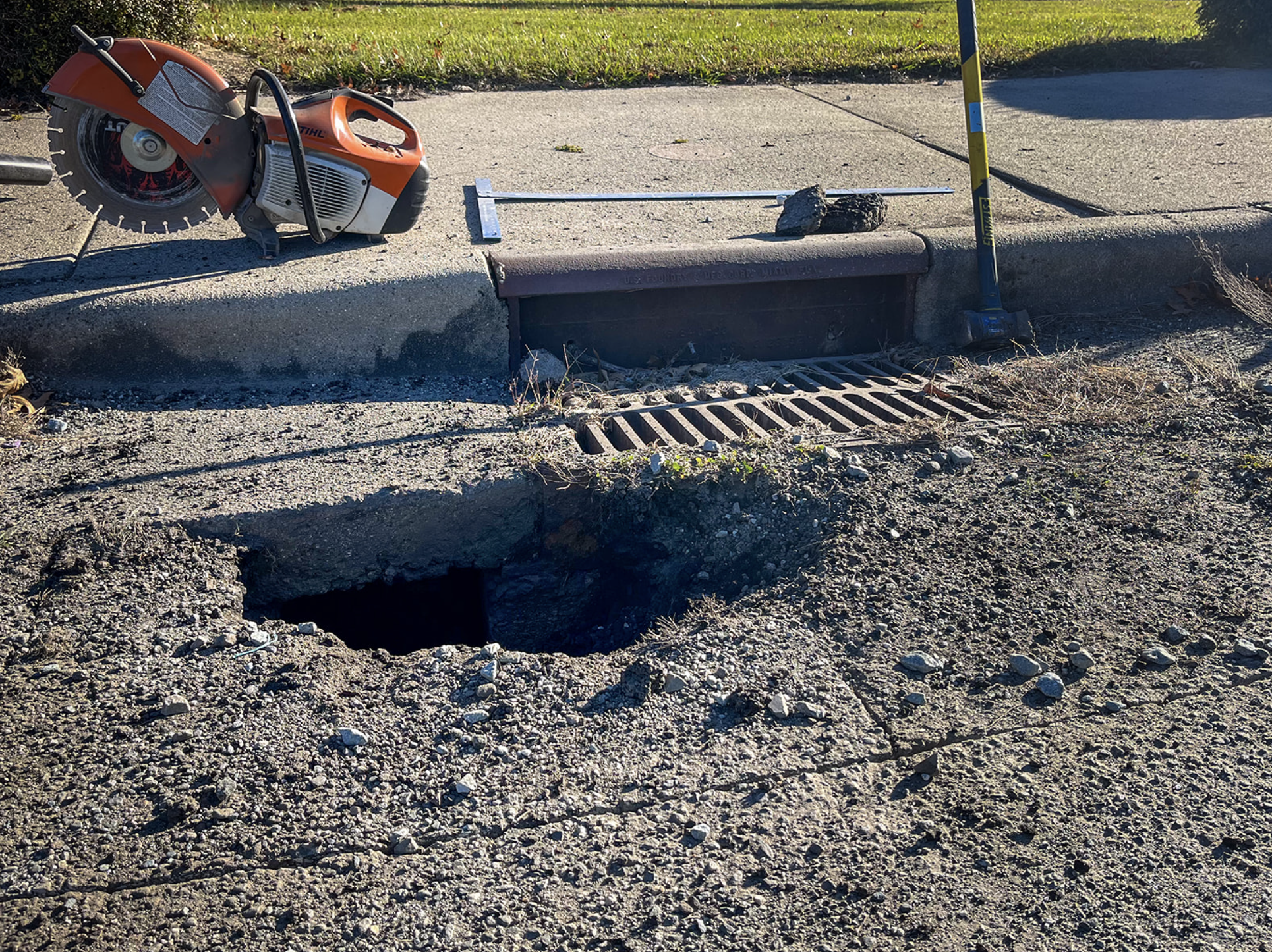 A storm drain on a street curb with a large hole and loose asphalt, a construction marker, a rubble stone, a metal T-shaped tool, a grated storm drain, and a power saw resting on the curb.