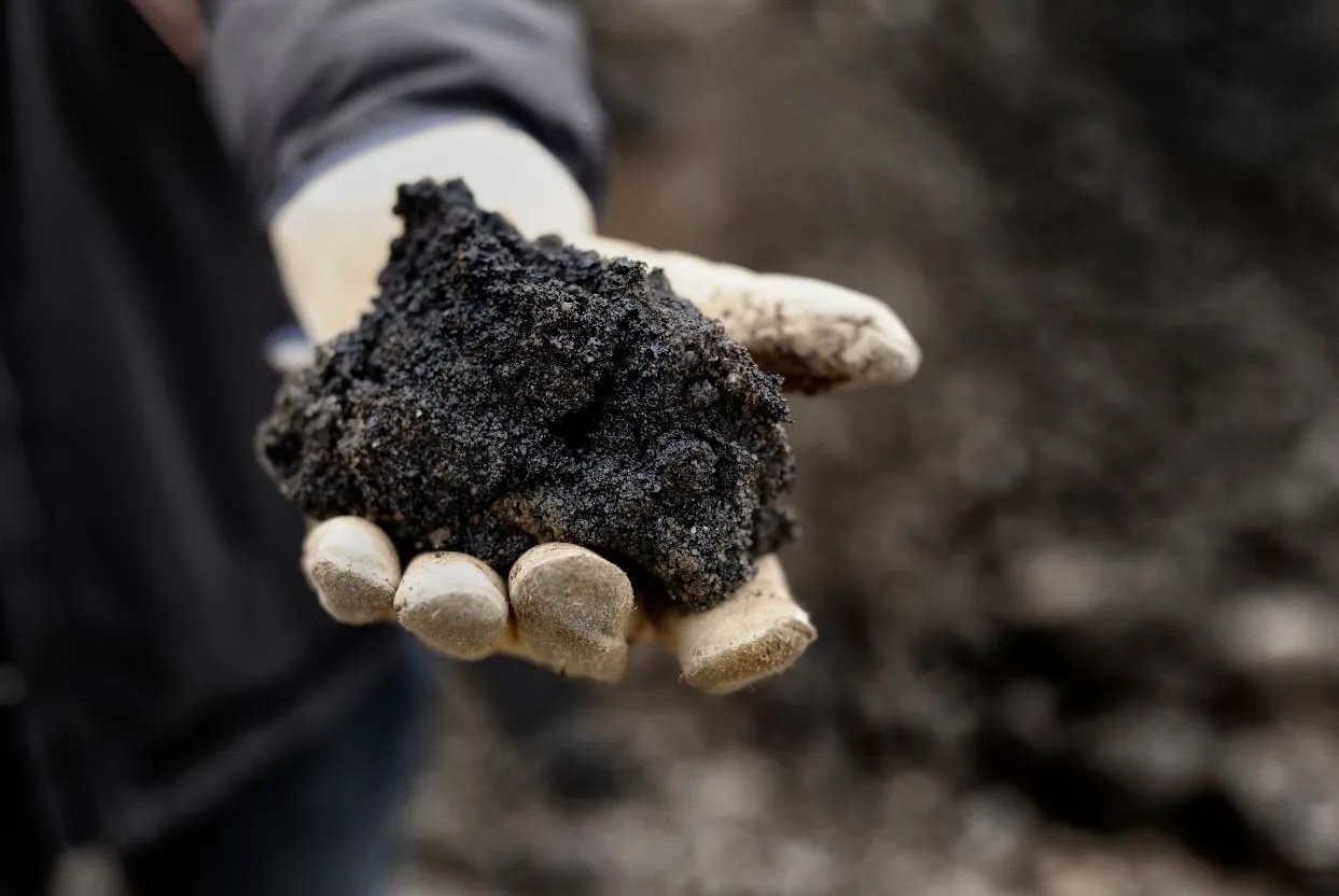 A gloved hand holding a chunk of raw asphalt millings, illustrating the raw material used in hot mix asphalt production.