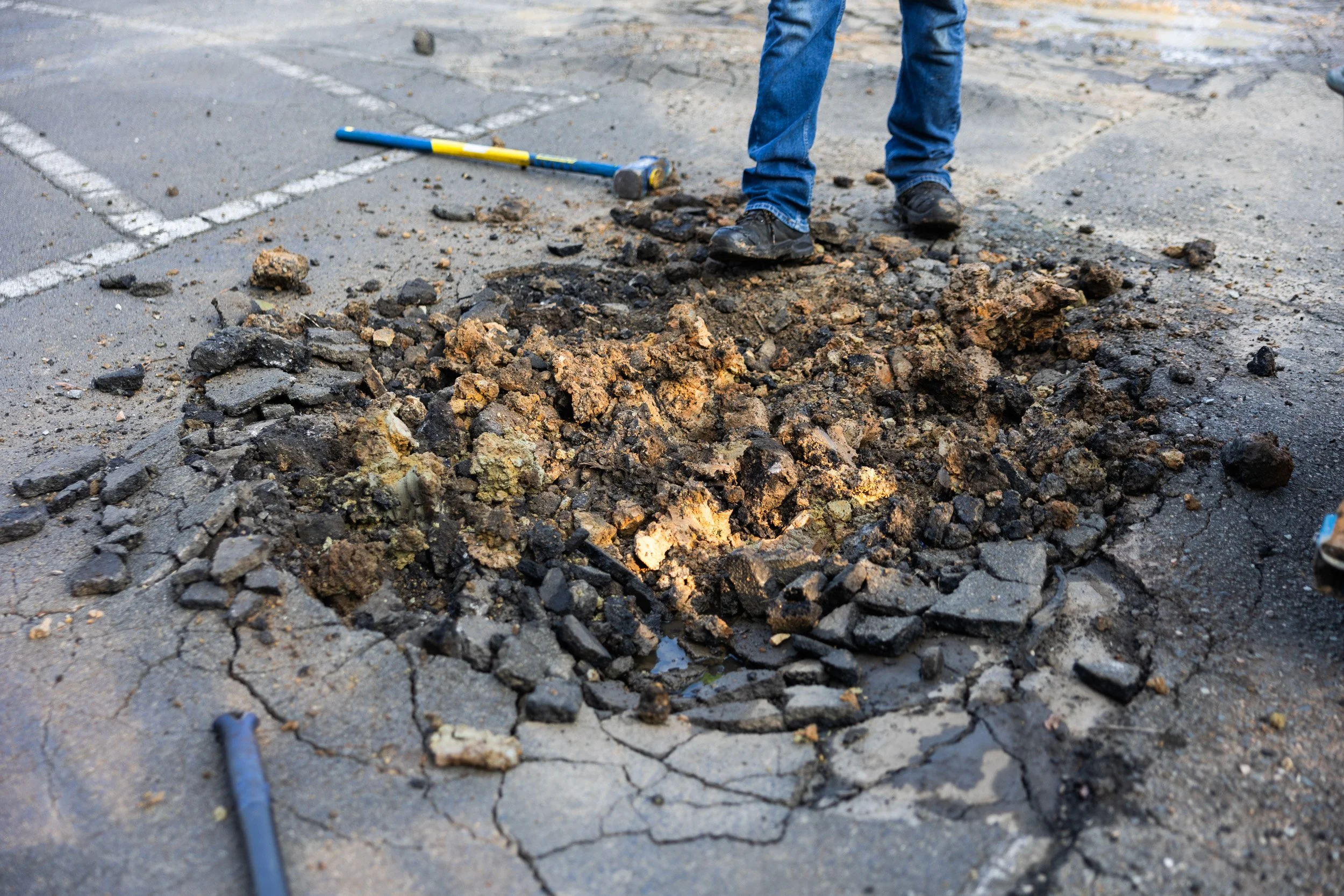 Person standing in a parking lot with a damaged pavement and broken asphalt, a hammer lying on the ground nearby.