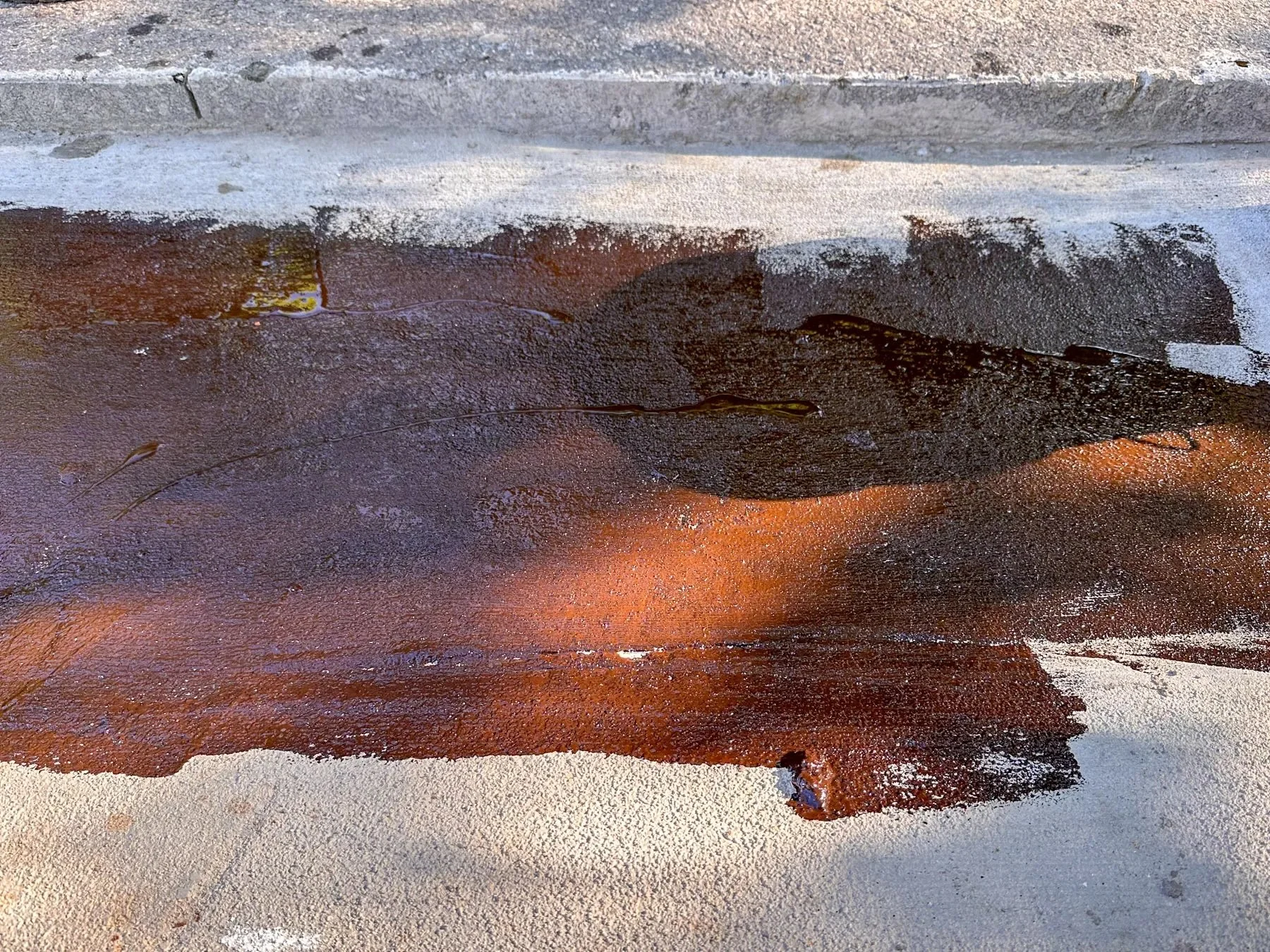 Close-up of reddish-brown asphalt emulsion tack coat freshly applied to a driveway apron next to a concrete curb, Durham, NC.