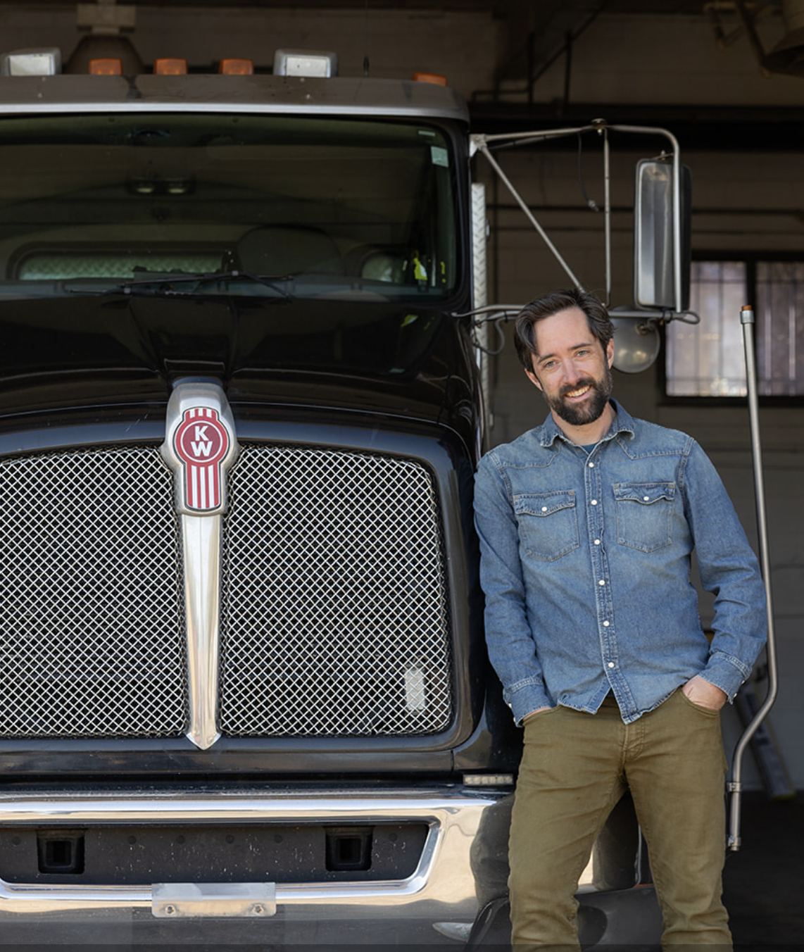 A man with a beard and dark hair, wearing a denim jacket and brown pants, standing and smiling next to a large black truck with a red and white Kenworth Trucks logo on the front grill, inside a garage.