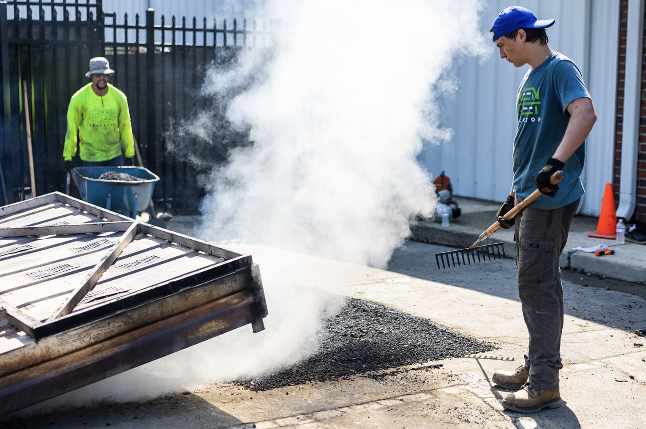 Two workers, one with a rake and the other with a shovel, are patching a pothole with an infrared repair truck near a white building. Steam rises from the fresh repair, and there is an orange cone in the background.