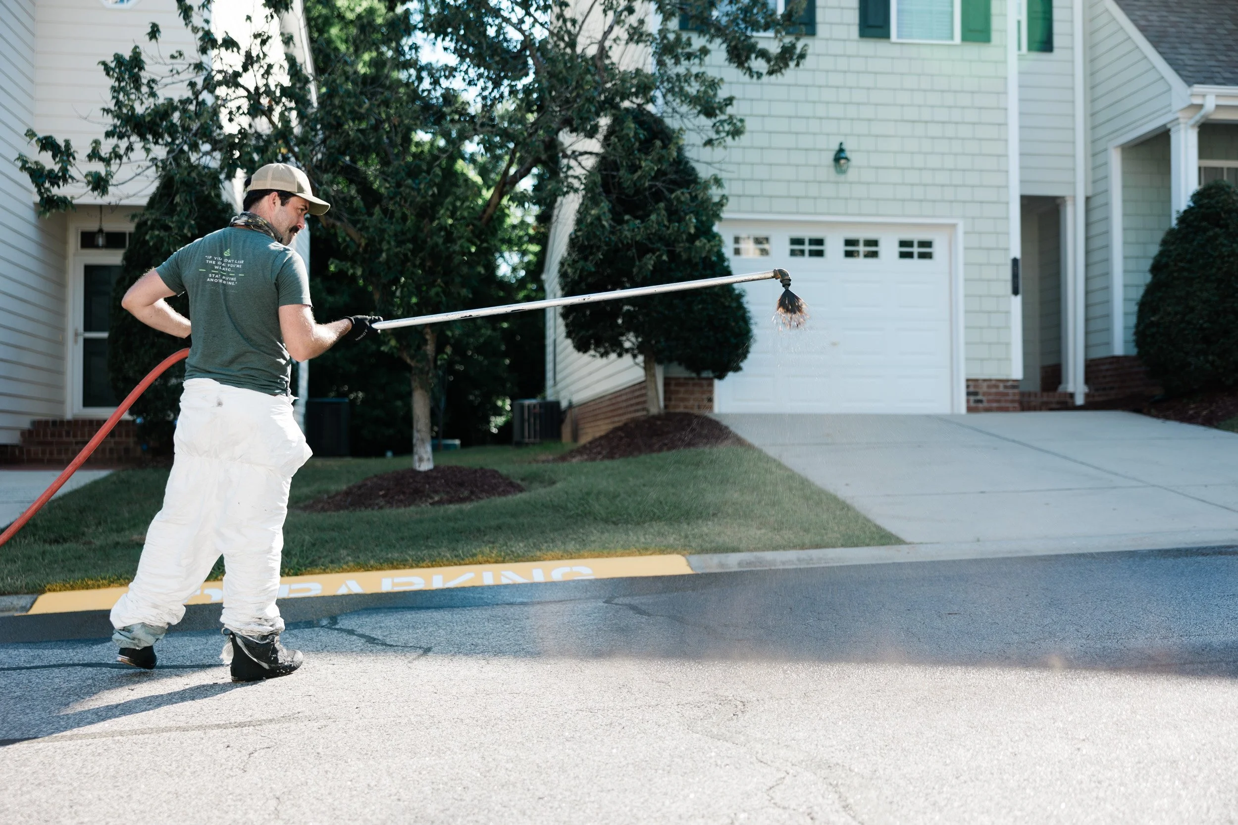 A man wearing a cap, t-shirt, and white pants is sealcoating asphalt with a wand sprayer outside a townhouse on a sunny day in Durham, NC.