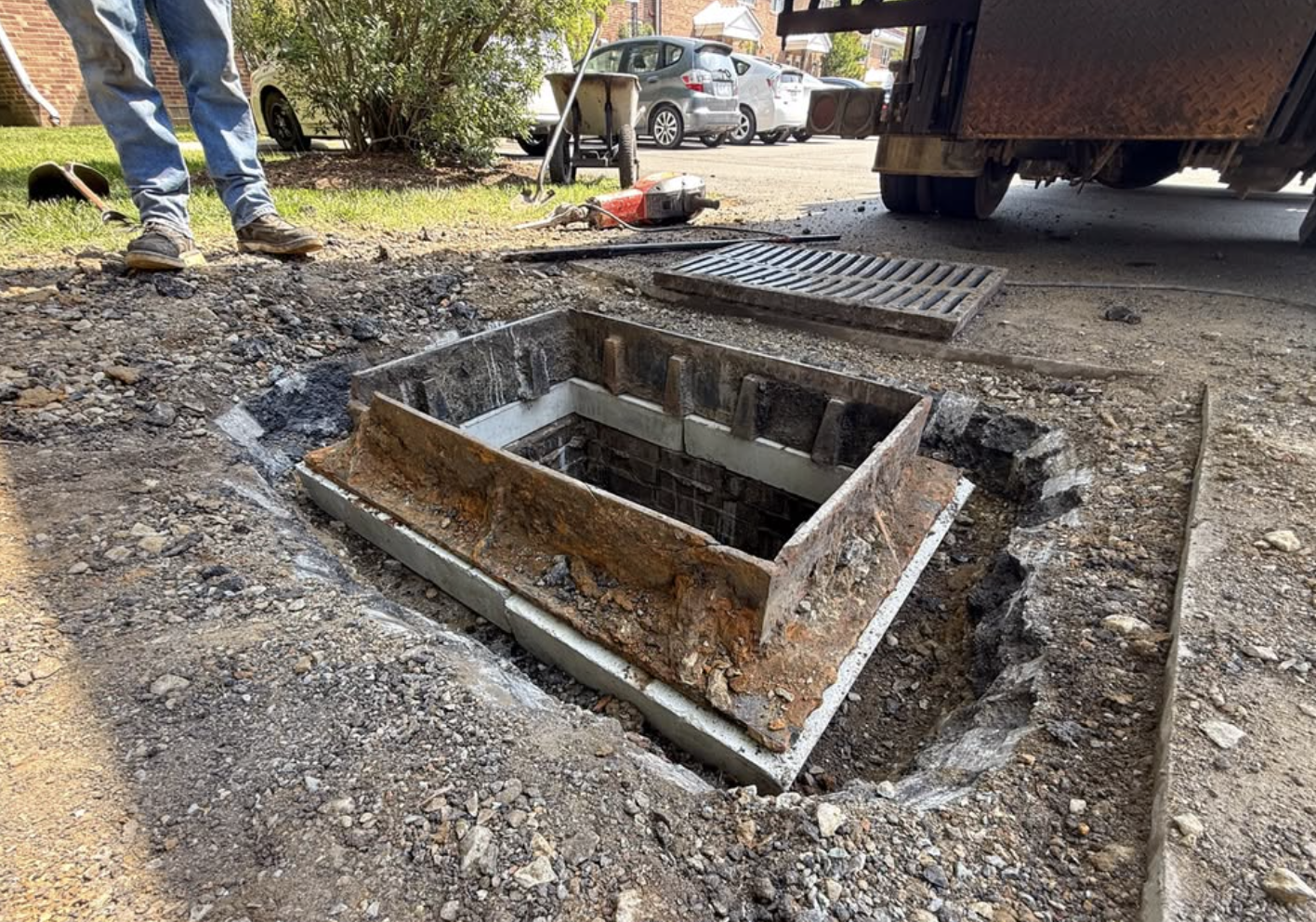 Open storm drain with a rusted metal frame, surrounded by dirt and gravel, in a residential area with parked cars and greenery in the background.