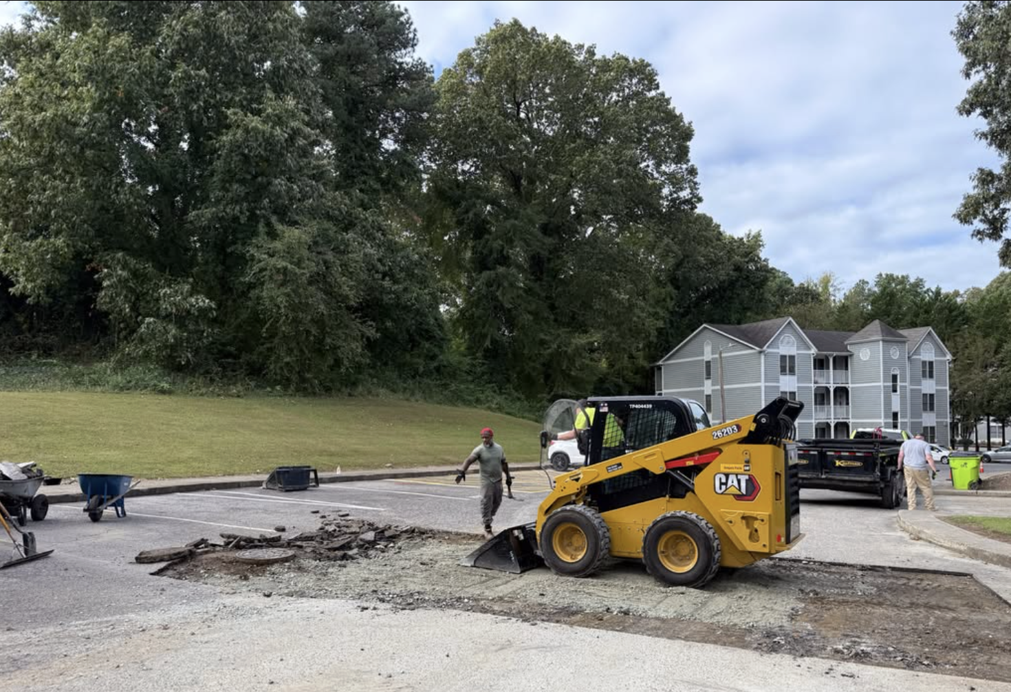 Construction workers repair a road in Durham, NC with a small yellow Caterpillar skid-steer with a milling attachment. Workers are visible wearing safety gear.