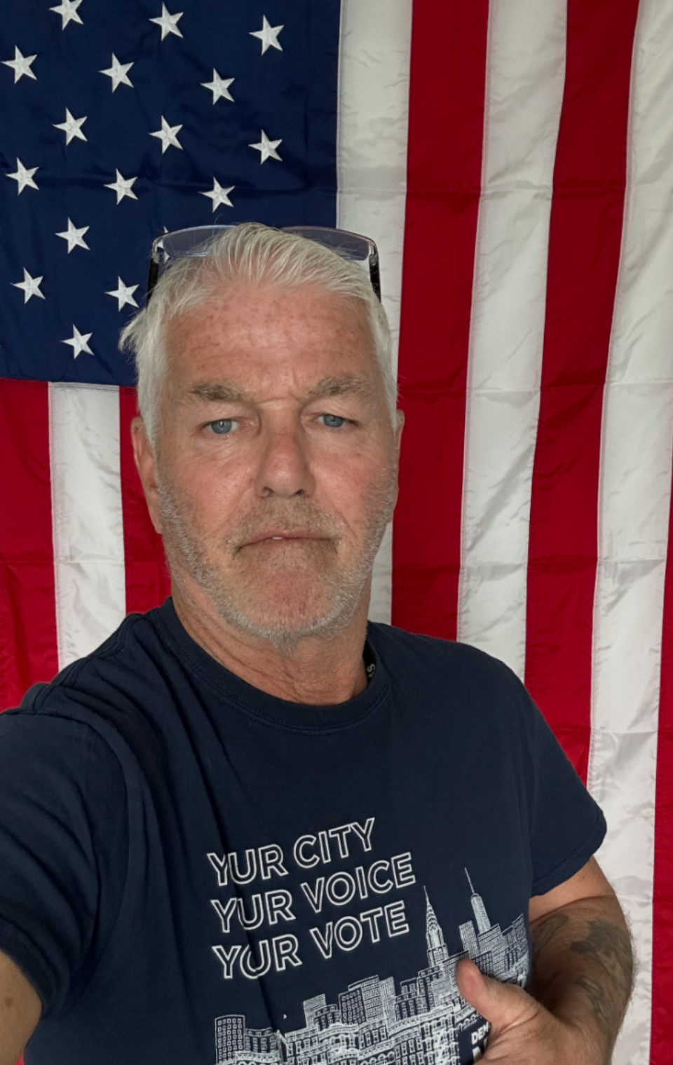 George Marrsh, a man with short, light-colored hair and a beard taking a selfie in front of an American flag. He is wearing a navy blue T-shirt with a cityscape graphic and the words 'YUR CITY YUR VOICE YUR VOTE.'
