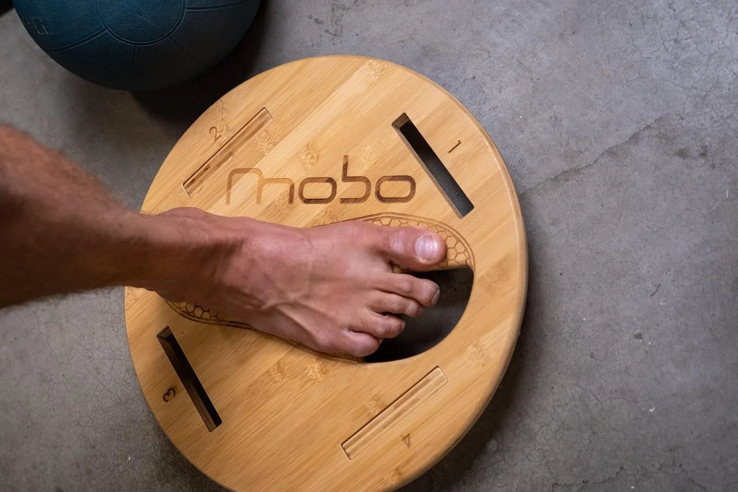 A person placing their foot on a wooden balance board with the word 'nobo' engraved on it, on a concrete floor next to a black medicine ball.