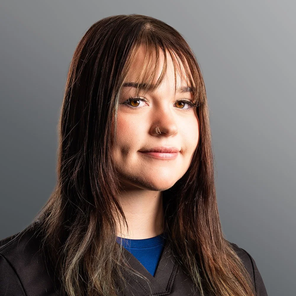 A young woman with brown hair, hazel eyes, and a nose piercing, wearing a black top, against a neutral gray background.