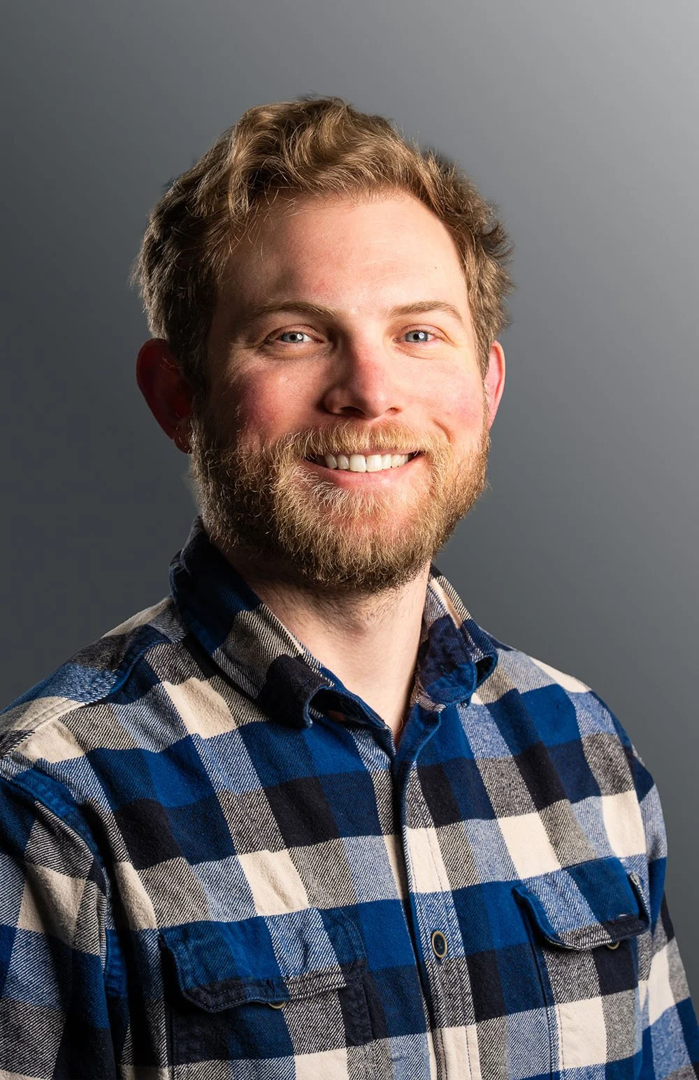 A smiling man with red hair and beard wearing a blue and white plaid shirt against a gray background.