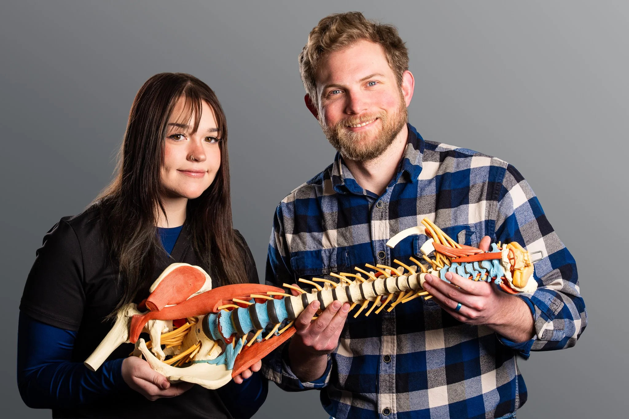 A man and woman holding a model of a human spine against a gray background.