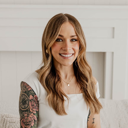 Young woman with long wavy hair smiling, wearing a white top, with tattooed arms, indoors against a white wall.