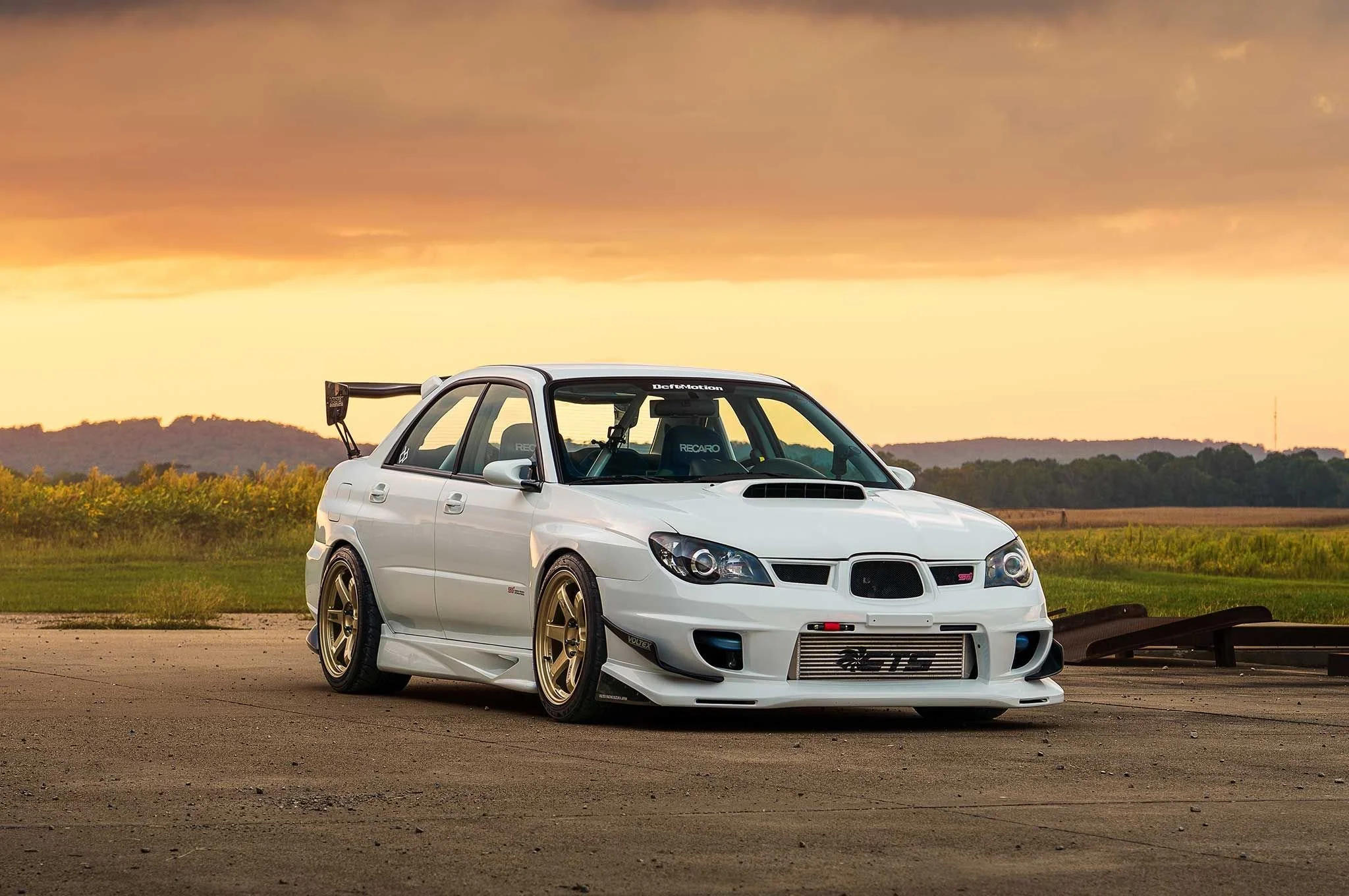 A white modified Subaru WRX STI with gold wheels parked on an open lot at sunset, with a scenic landscape and dark clouds in the sky.