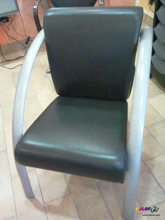 A black padded chair with white metal armrests and legs, sitting on a tiled floor in an indoor setting.