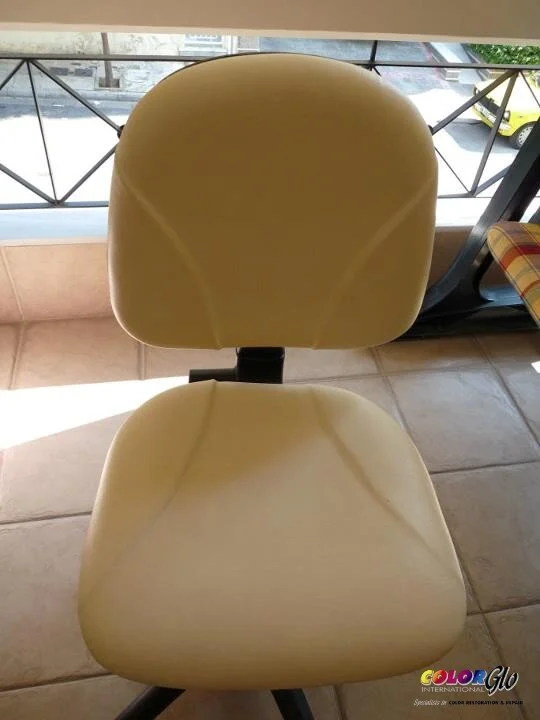 A beige office chair with a rounded backrest and seat, placed on a tiled floor in front of a window with metal railings and a view of the street outside.