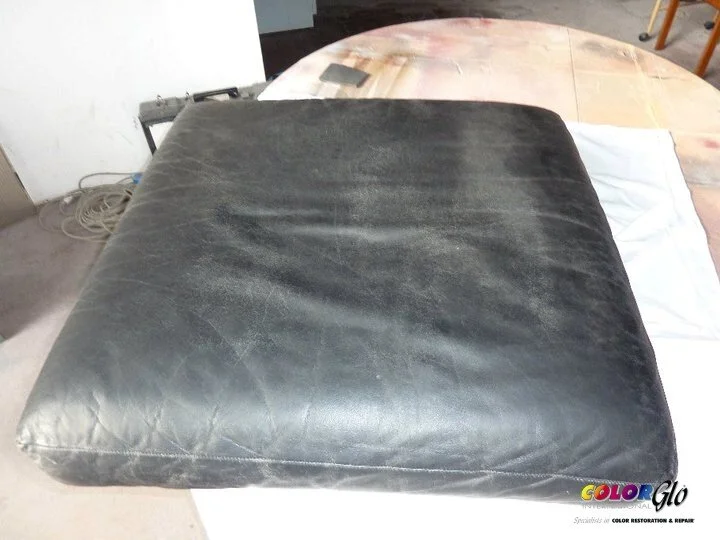 A black leather cushion or seat pad placed on a white surface in a workshop or repair shop setting.