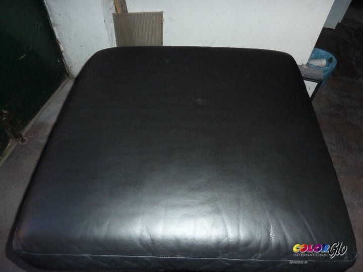 A black leather ottoman or cushion in a room with a dark floor and a white wall, surrounded by some household items.