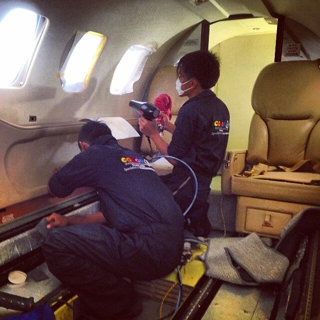Two workers in uniforms with a TV5 logo cleaning inside an airplane, one is using a cleaning tool and the other is with a handheld device, both wearing masks.