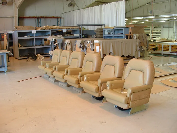 Six beige leather seats in a factory or warehouse setting, with shelves and workspaces in the background.