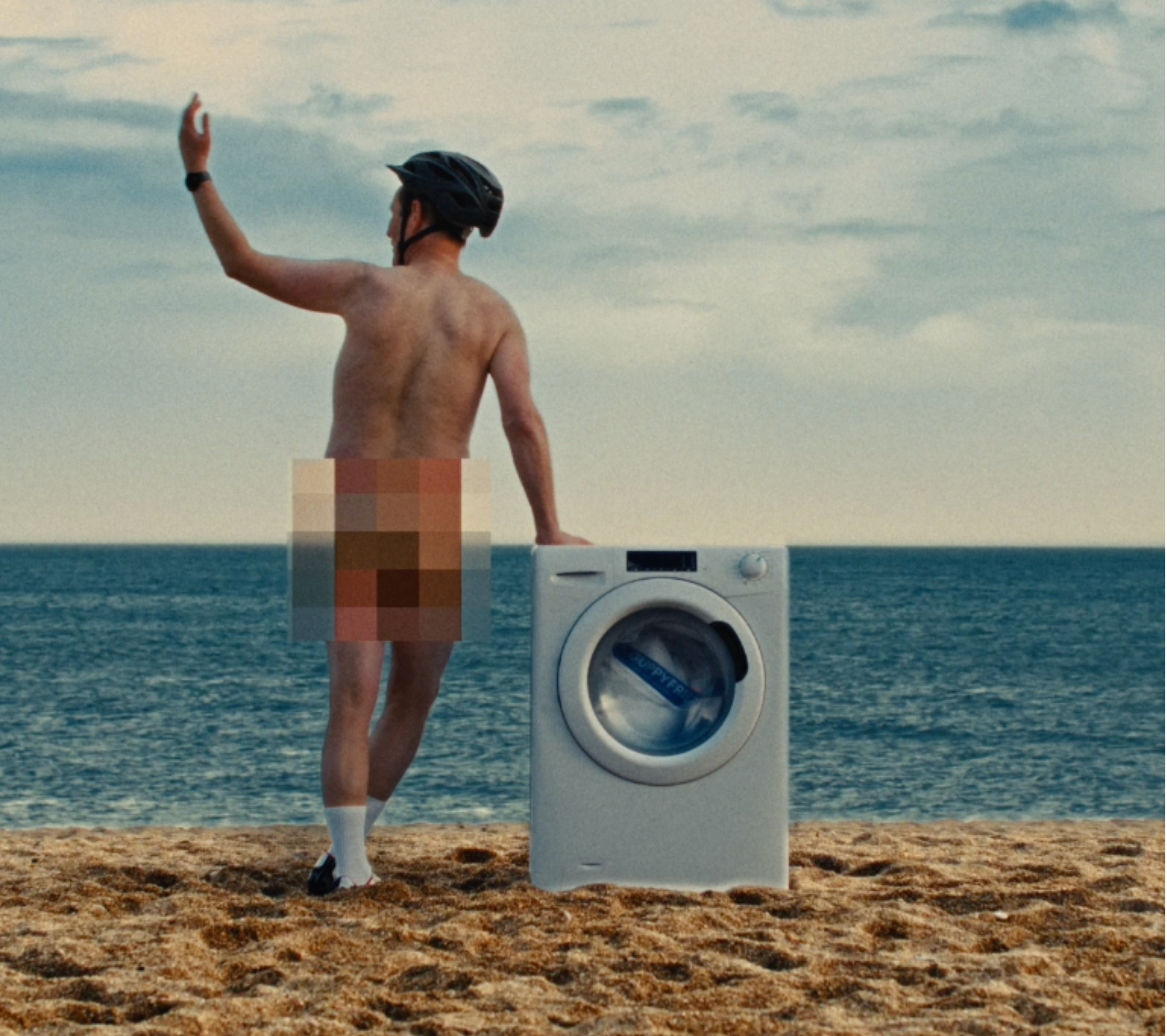 Man standing in fronf of the ocean, with a washing machine besides him.