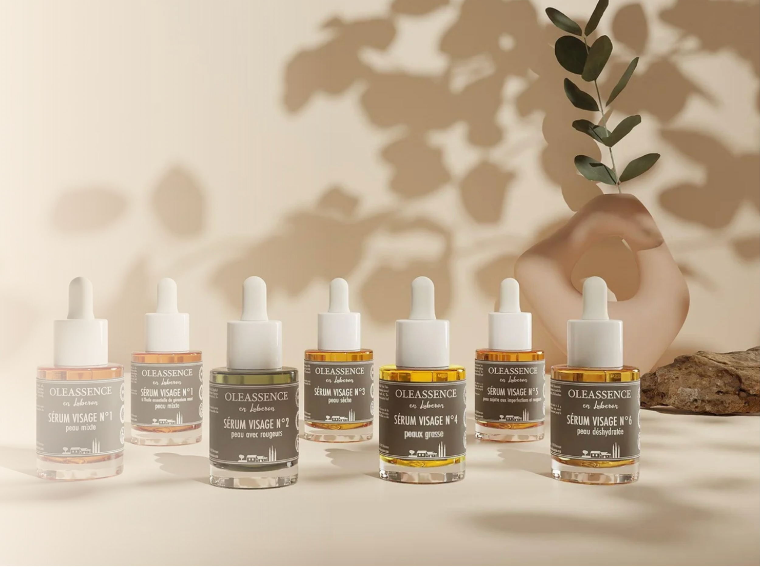 A row of small bottles of Oleassence skincare serums with droppers on a white surface, with a hand holding a small leafy branch in front of a backdrop casting shadows.
