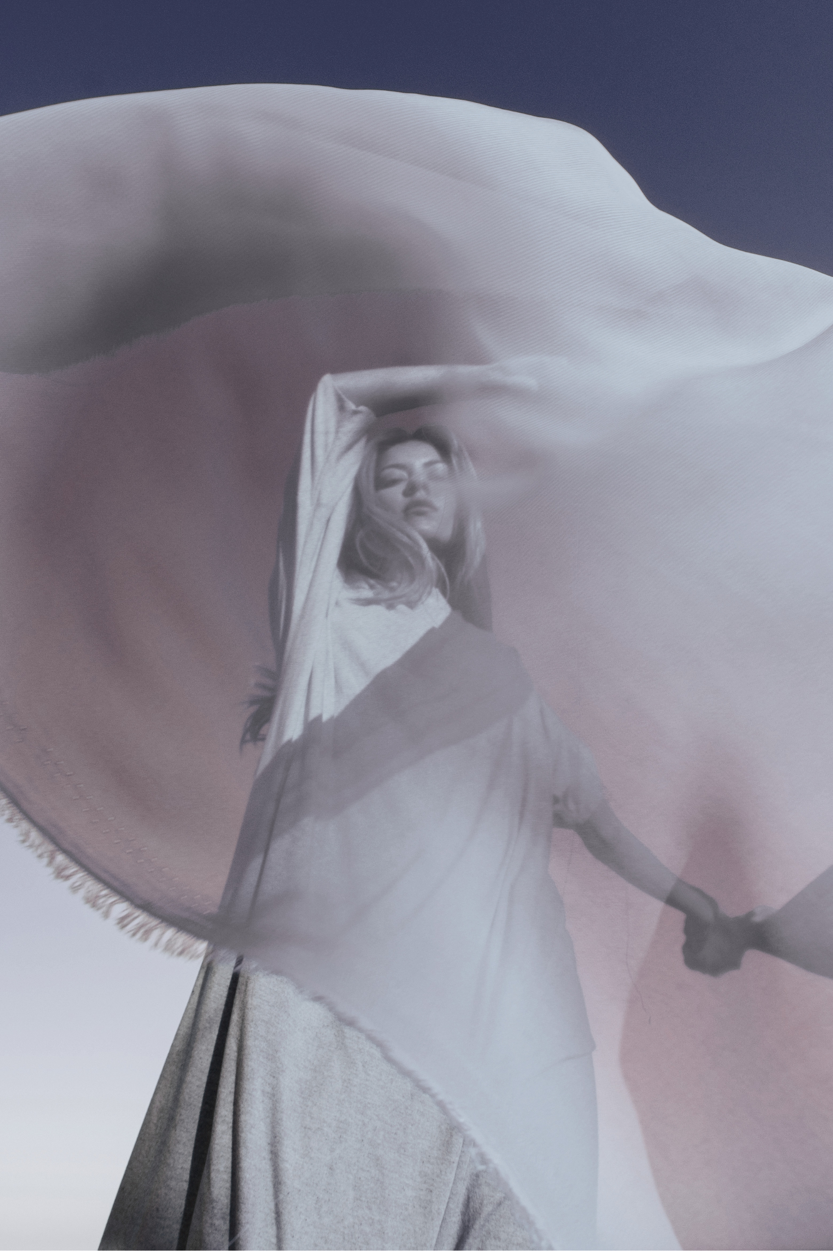 A woman holding a white parasol while standing outdoors, with her eyes closed and one arm resting on her head, under a clear blue sky.