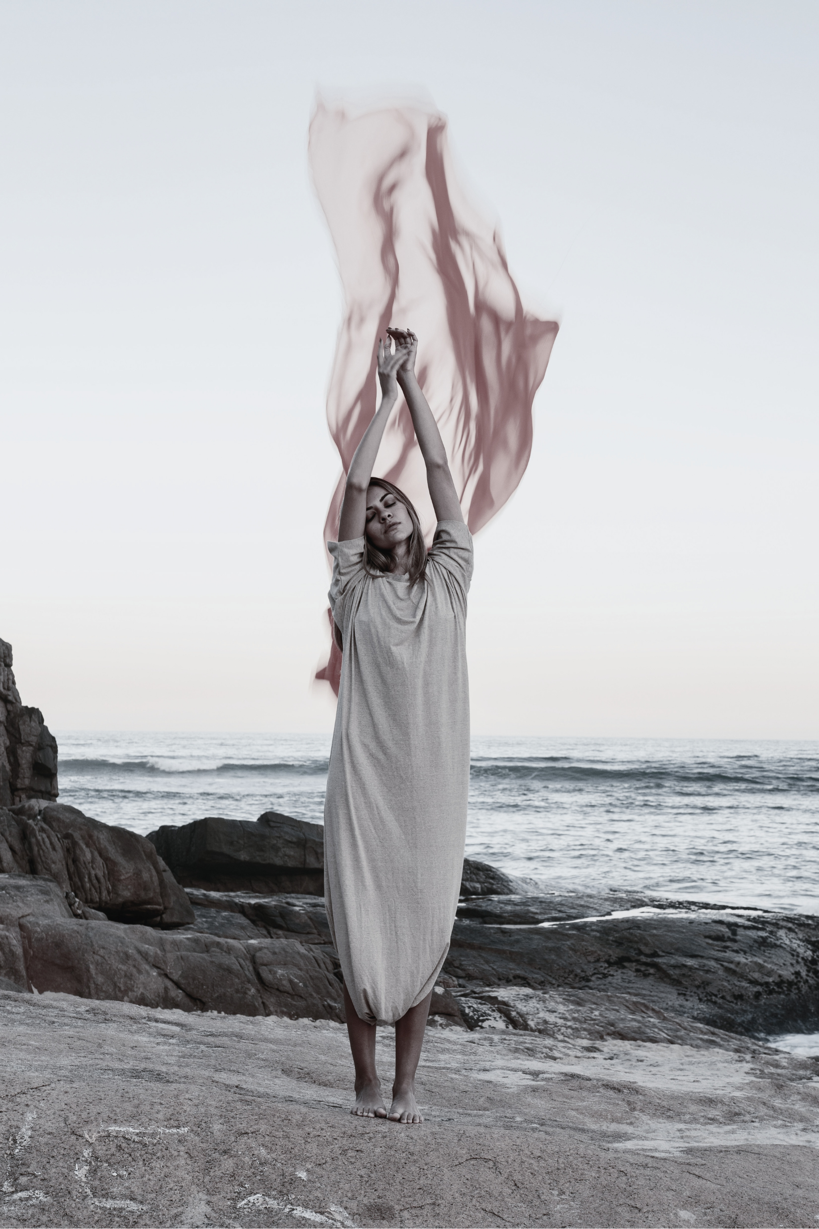 A woman standing barefoot on a beach with rocks, holding a flowing pink fabric above her head as it billows in the wind, with the ocean and cloudy sky in the background.