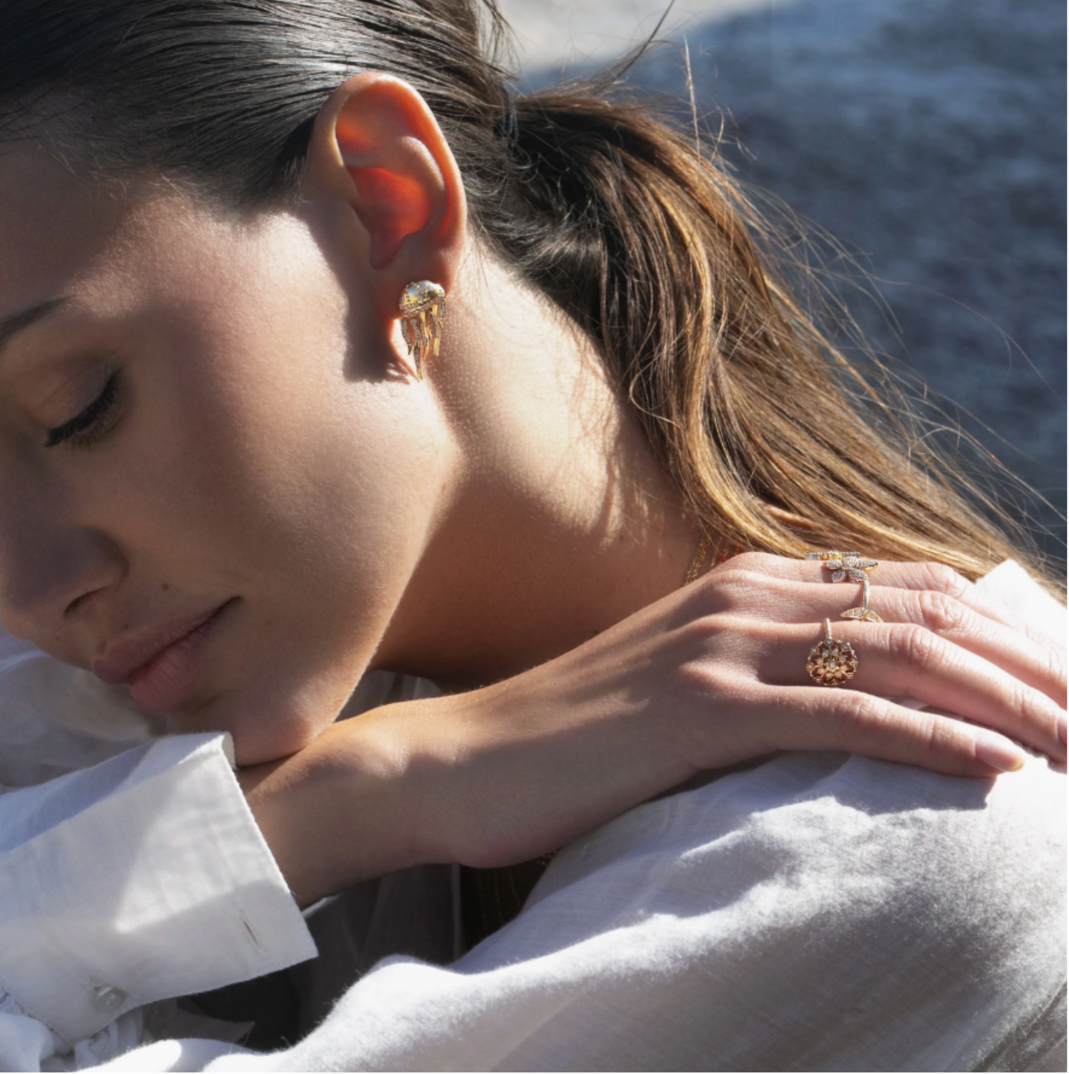 Close-up of a woman wearing jewelry including earrings, rings, and a necklace, with her face partially visible, shot outdoors near water.