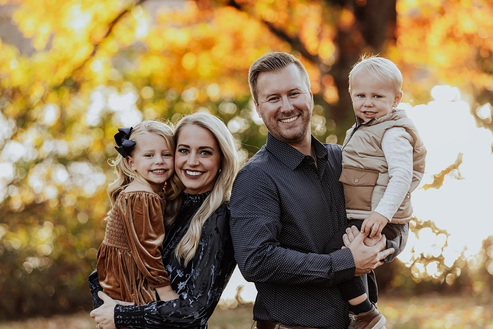 A family of four smiling outdoors during fall, with autumn leaves in the background, including a woman, a man holding a young boy, and a girl with a bow in her hair
