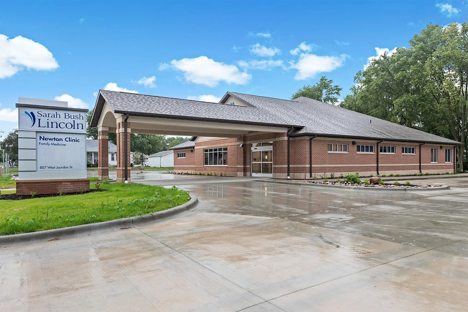 Exterior view of a brick medical clinic building with a large sign that reads 'Sarah Bush Lincoln Newton Clinic Family Medicine, 807 West Jourdan St.' The building has a parking lot, some greenery, and trees in the background. The sky is partly cloudy with some sunshine.