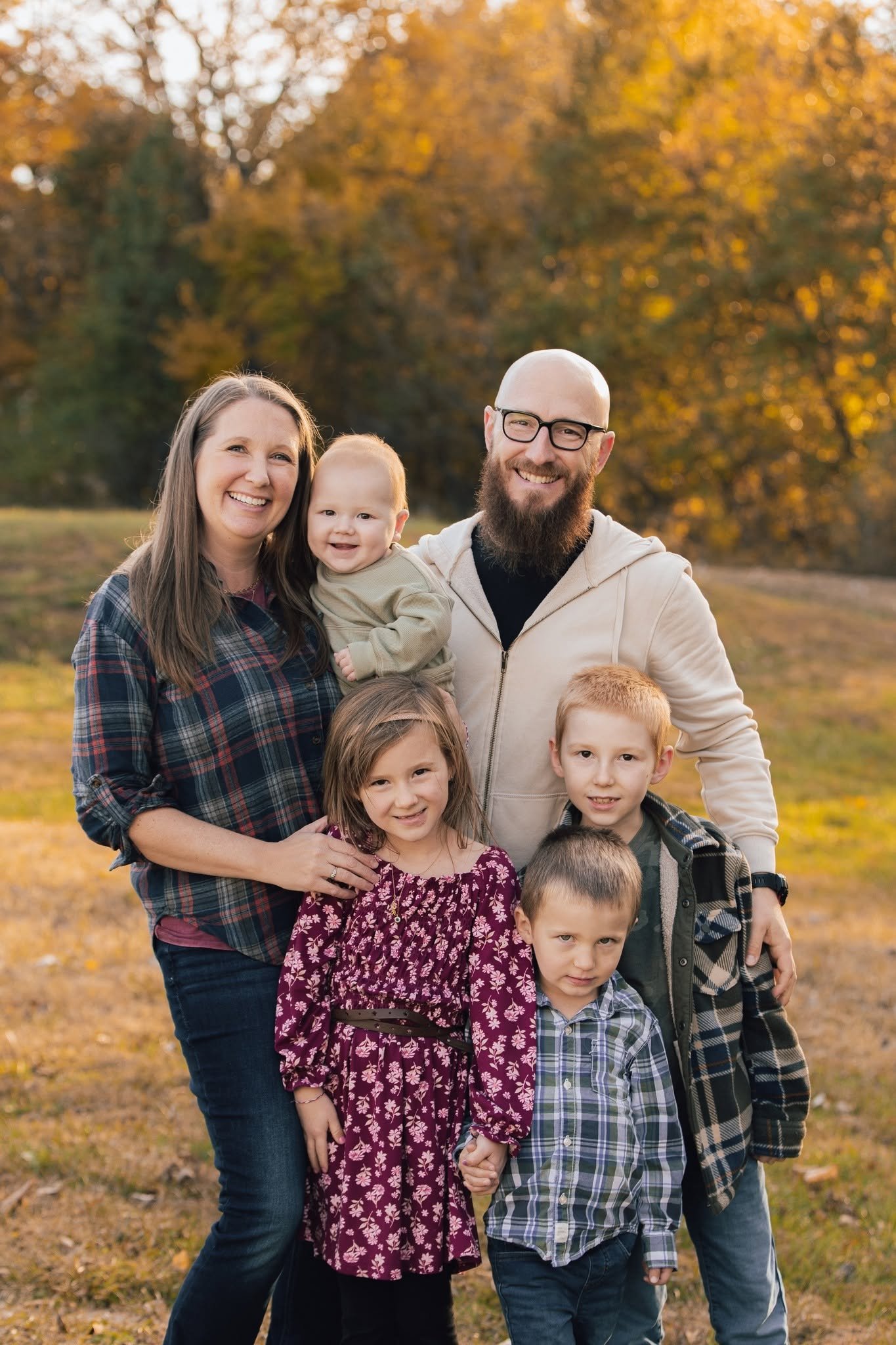 A smiling family of seven posing outdoors in a park during autumn, with trees with yellow leaves in the background.