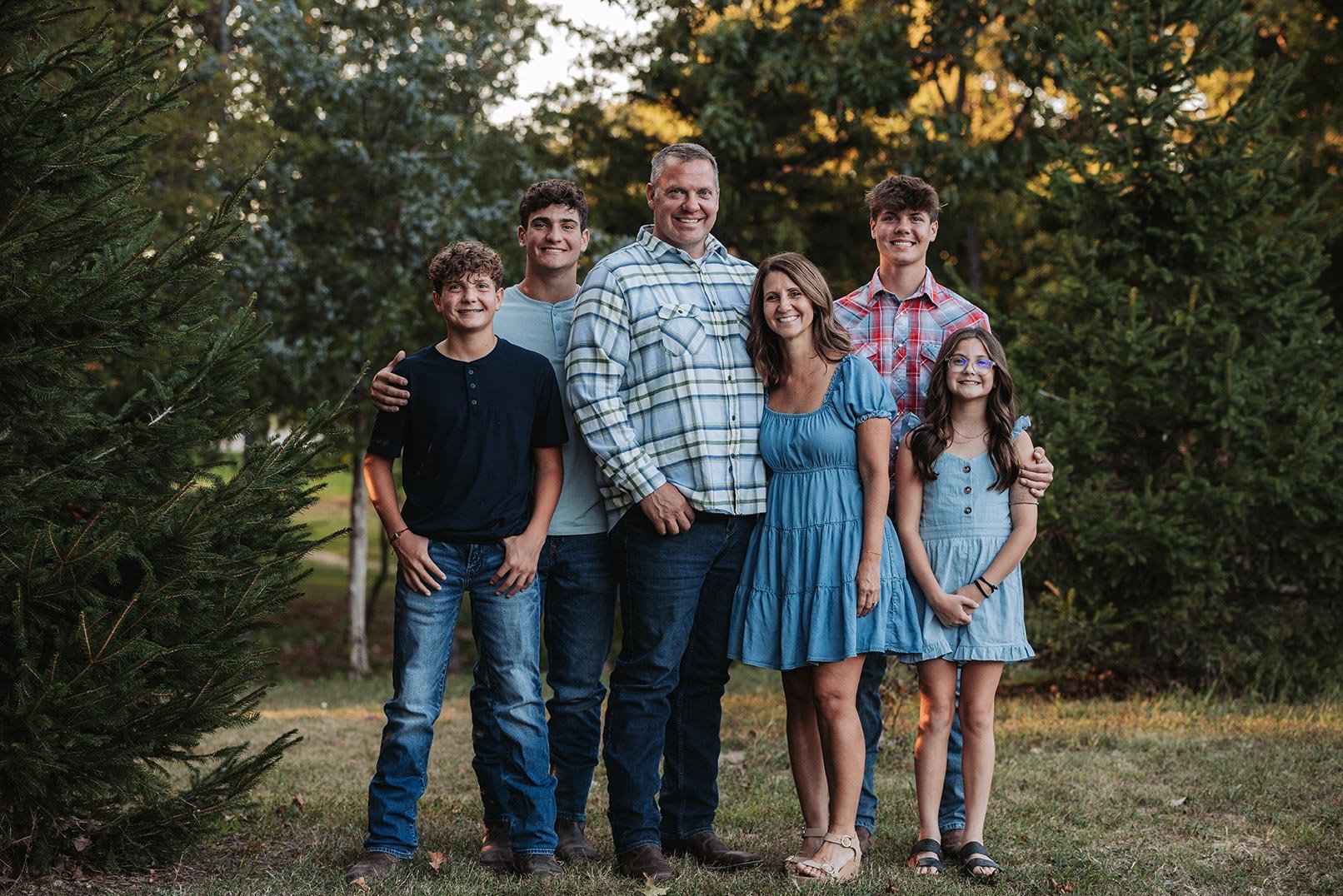 A family of six standing outdoors among trees at sunset, smiling for a group photo, with three males, two females, and a girl, wearing casual summer clothing.