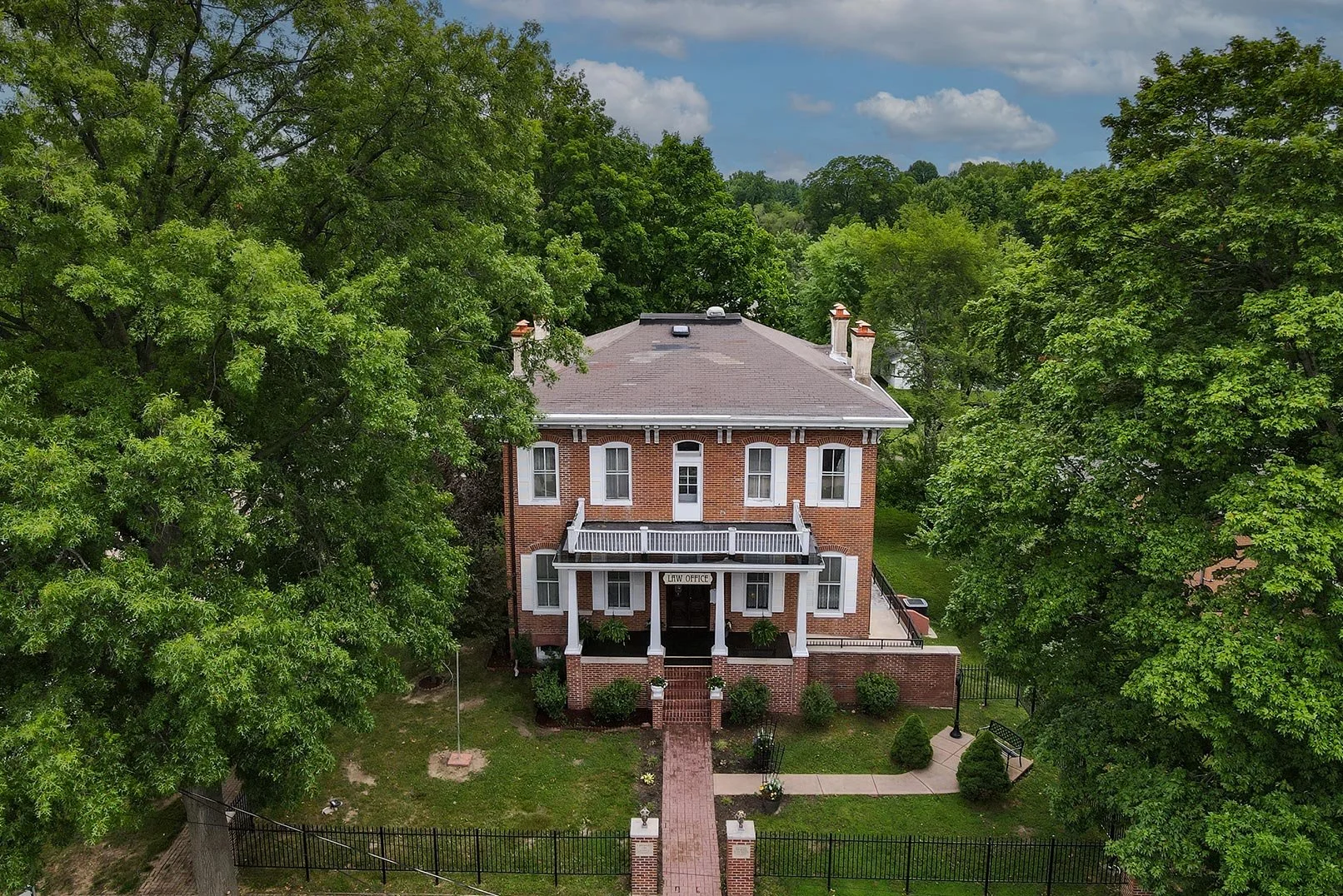 A large, two-story brick building with white trim labeled as a law office, surrounded by green trees, with a brick walkway leading to the entrance, and a black metal fence enclosing the property under a partly cloudy sky.