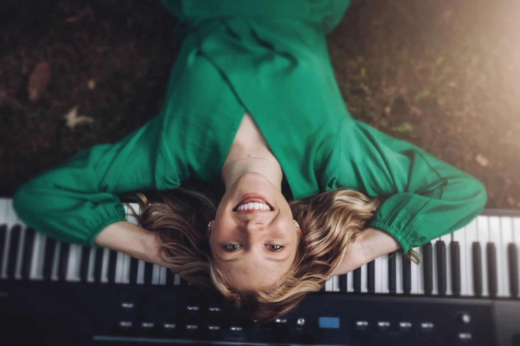 A woman lying on her back on a piano, smiling at the camera with her hands behind her head, wearing a green blouse, with brown hair spread out on the piano keys.
