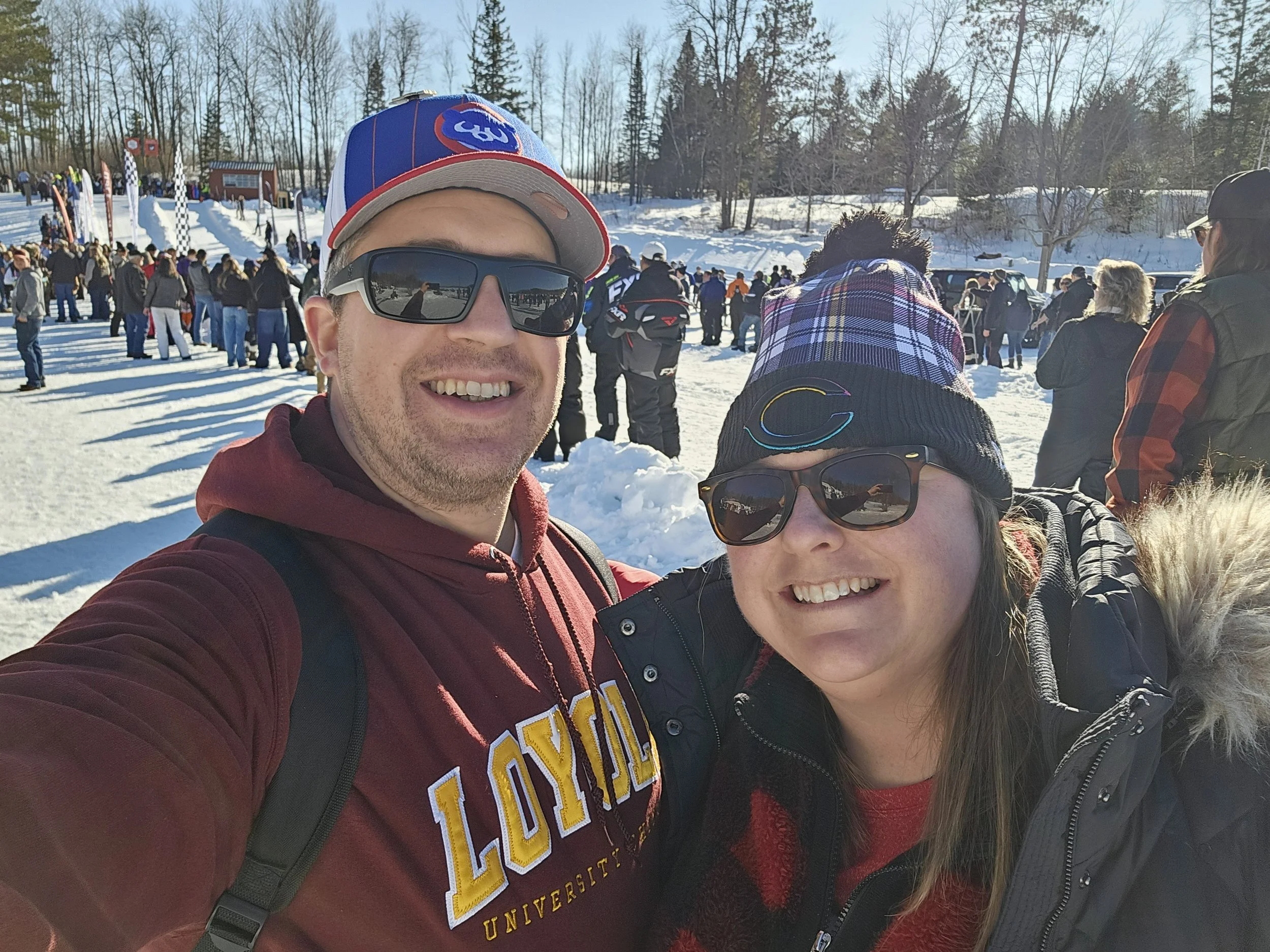 A man wearing sunglasses, a baseball cap, and a maroon hoodie smiling for a selfie at a snowy outdoor event. Behind him, a woman and a crowd of people gather near snow-covered land and trees, with a small building and flags visible in the background.