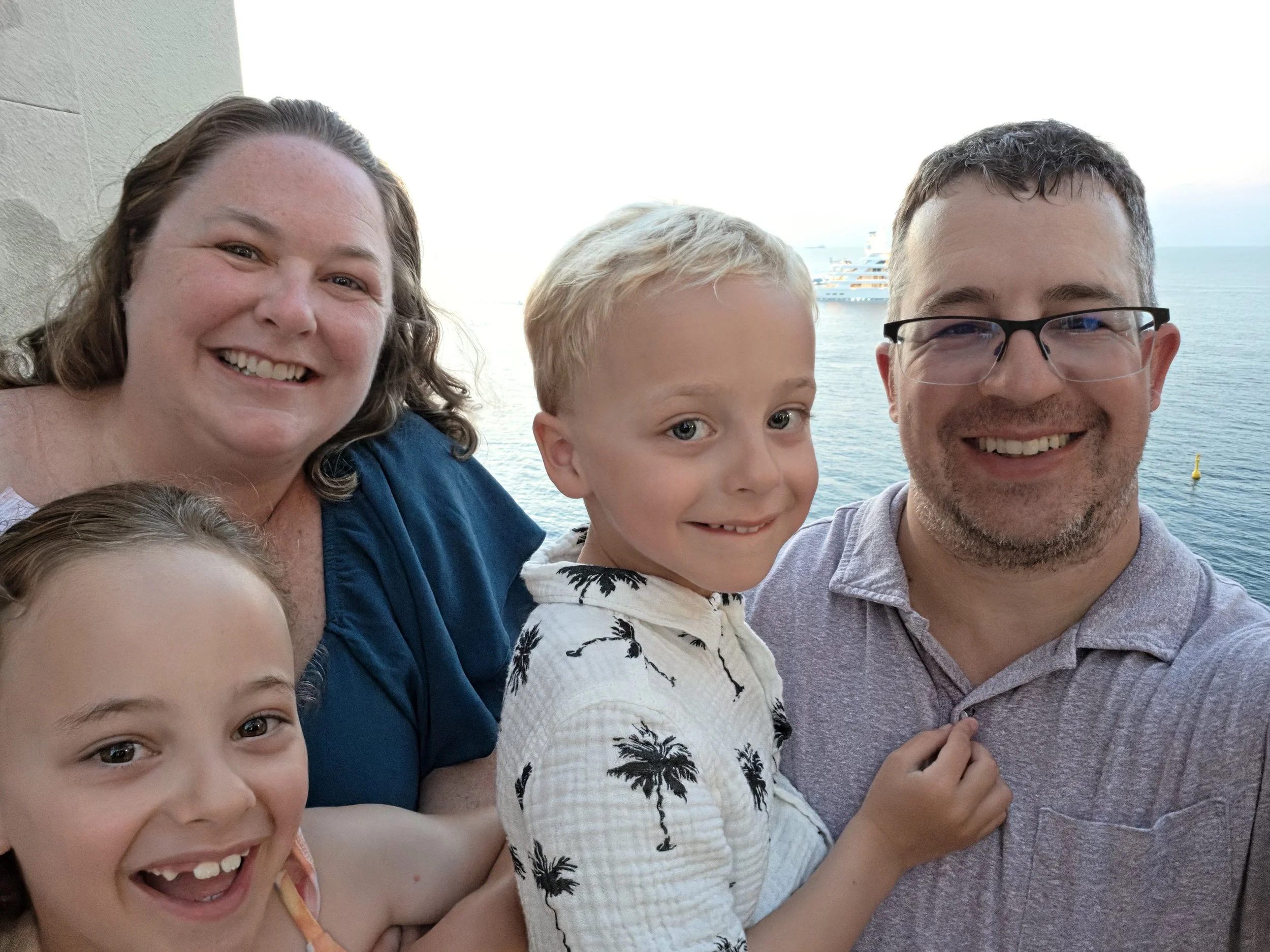 A family of five smiling for a selfie with the ocean and a cruise ship in the background.