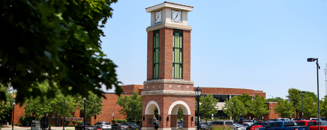 A brick clock tower with a white clock face and a peaked roof, located in a parking lot surrounded by trees and a brick building.