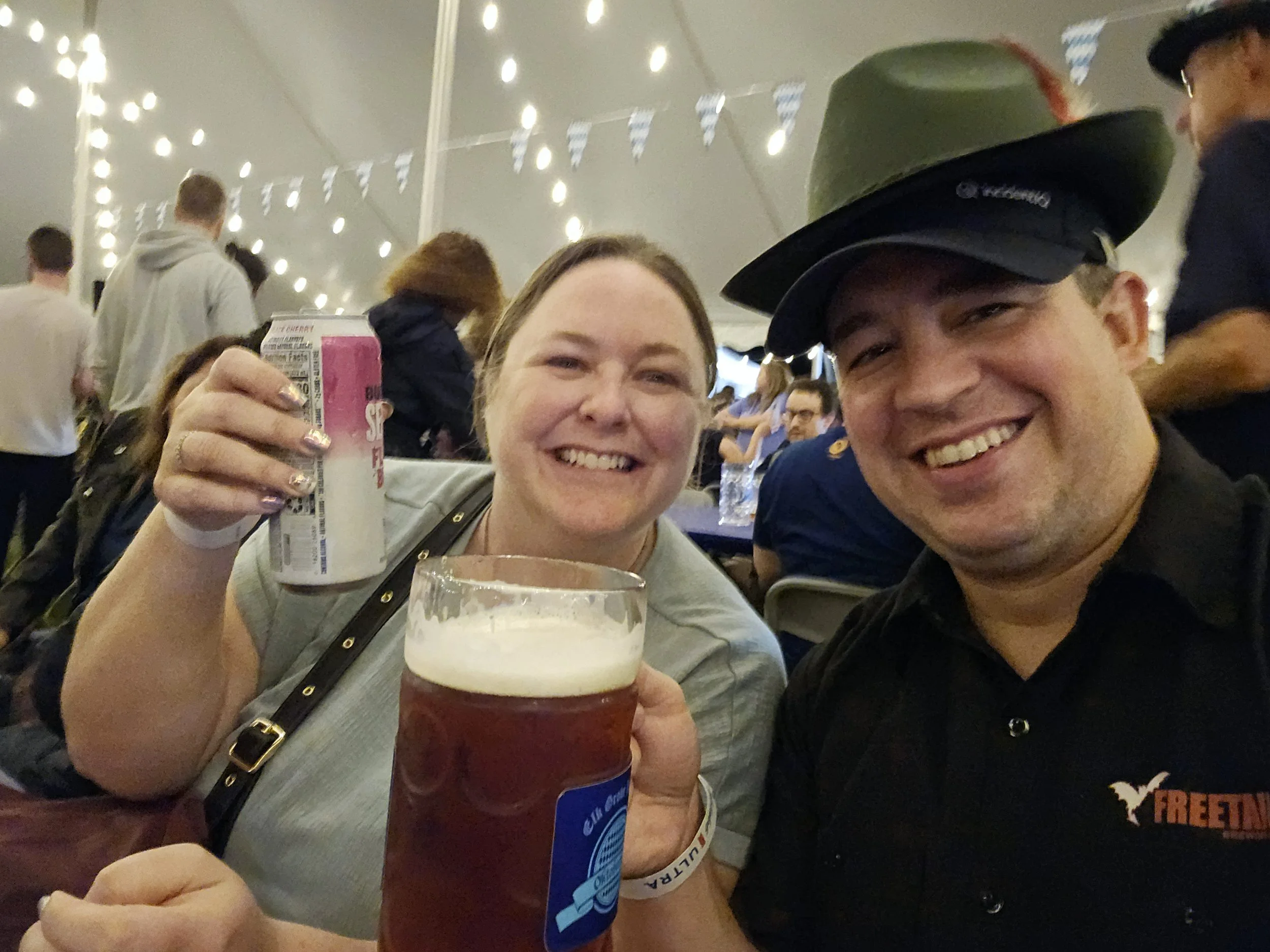 Two smiling women at a social gathering, one holding a pint of beer and the other holding a can, with people and string lights in the background.