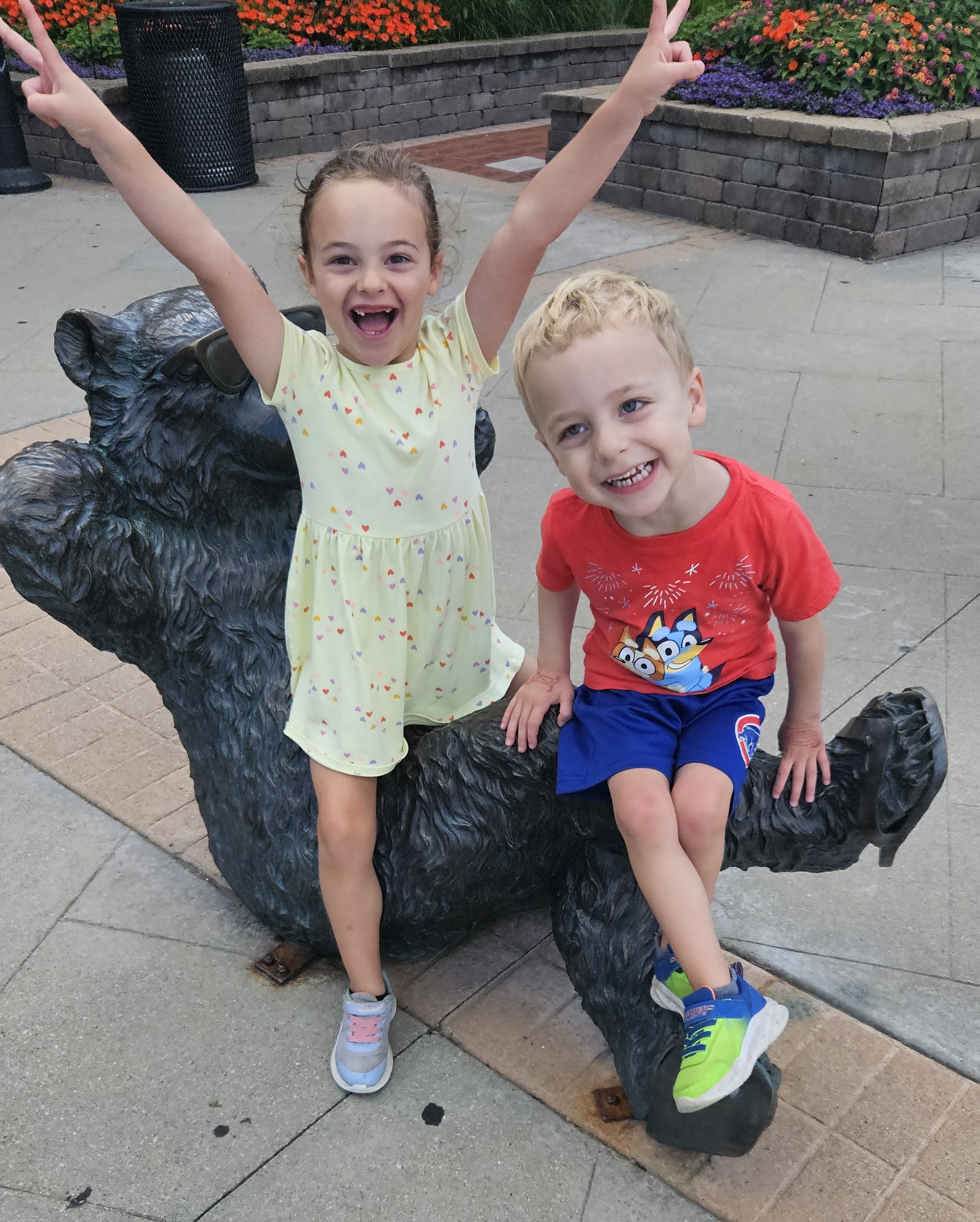 Two children, a girl and a boy, sitting on a bear-shaped sculpture in a park with flowers and brick planters in the background. The girl has brown hair, is wearing a yellow dress with colorful hearts, and is waving with both hands. The boy has blonde hair, is wearing a red shirt with cartoon characters and blue shorts, and is smiling at the camera.